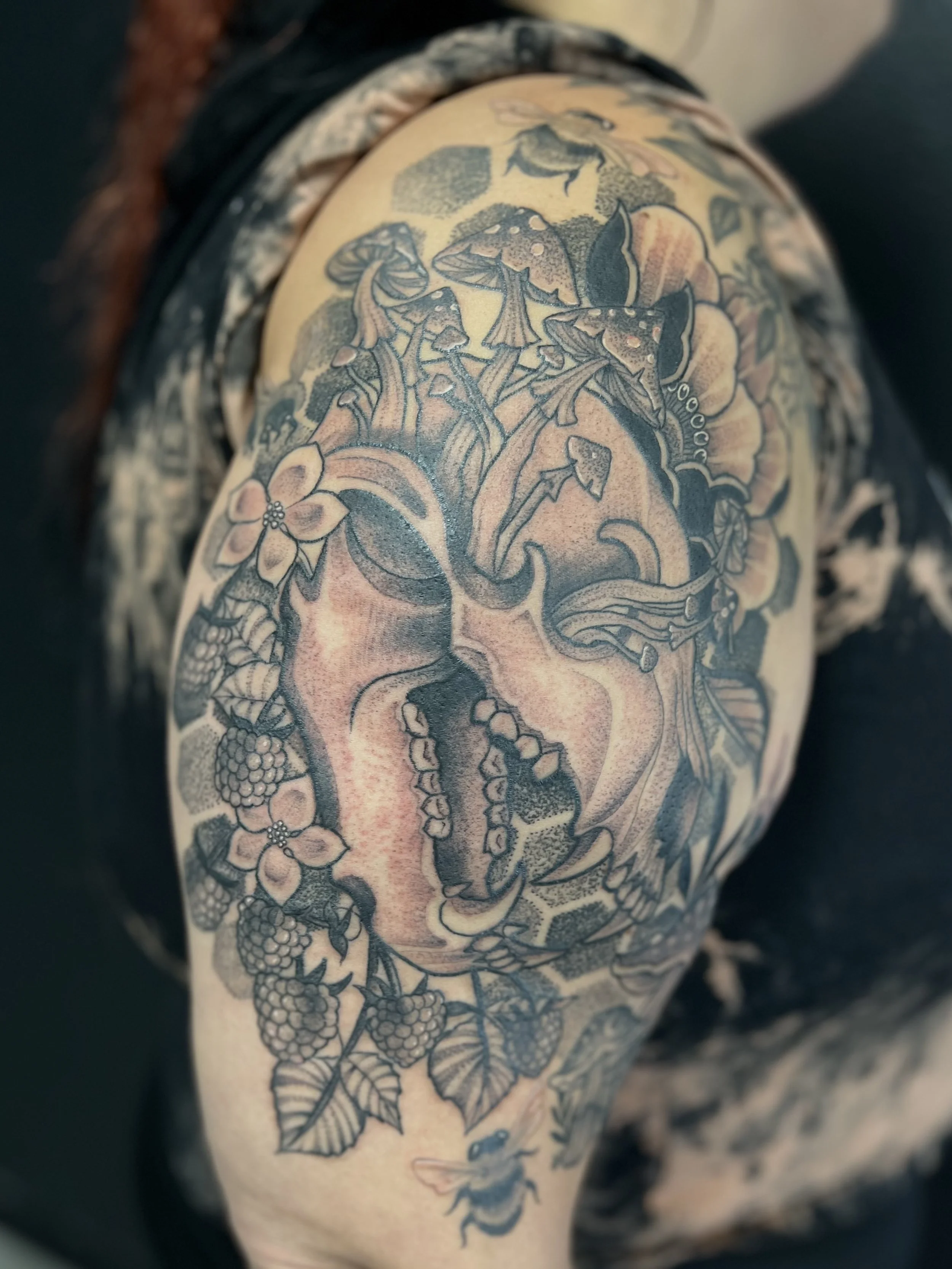 black and grey stipple style tattoo sleeve featuring a bear skull with mushrooms, flowers, berries, honeycomb and a bee on the shoulder.