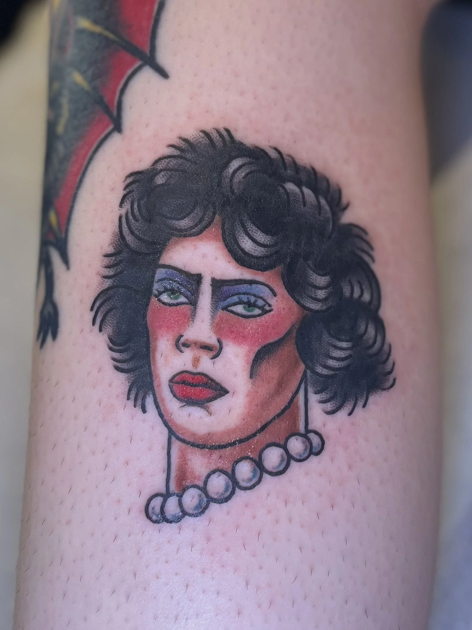 Tattoo of a rocky horror picture show character with dark wavy hair, bold makeup including blue eyeshadow, red blush, and red lipstick, wearing a pearl necklace.