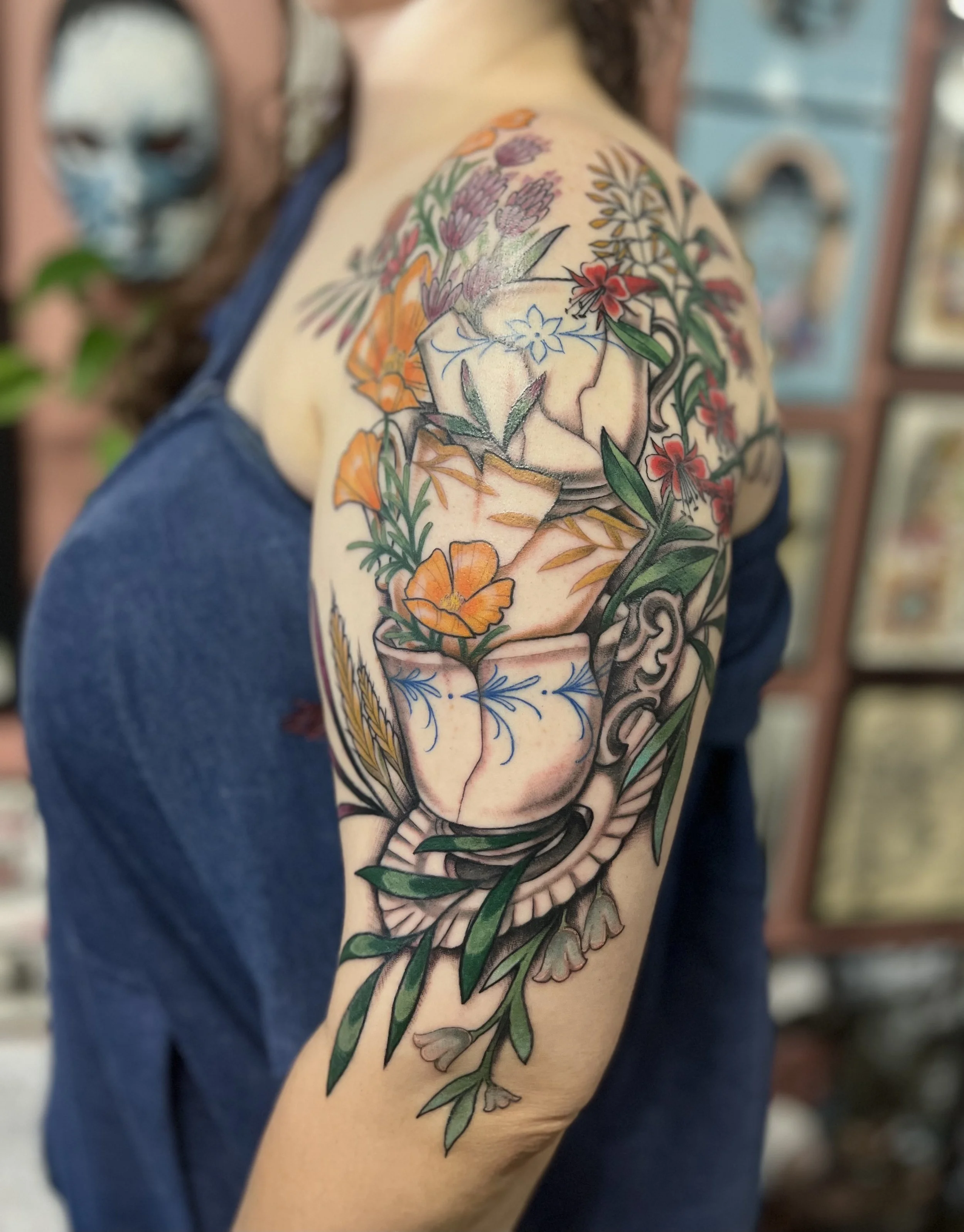 A  Neo traditional colorful tattoo on a person's upper arm and shoulder featuring a broken teacups with blooming flowers and foliage.