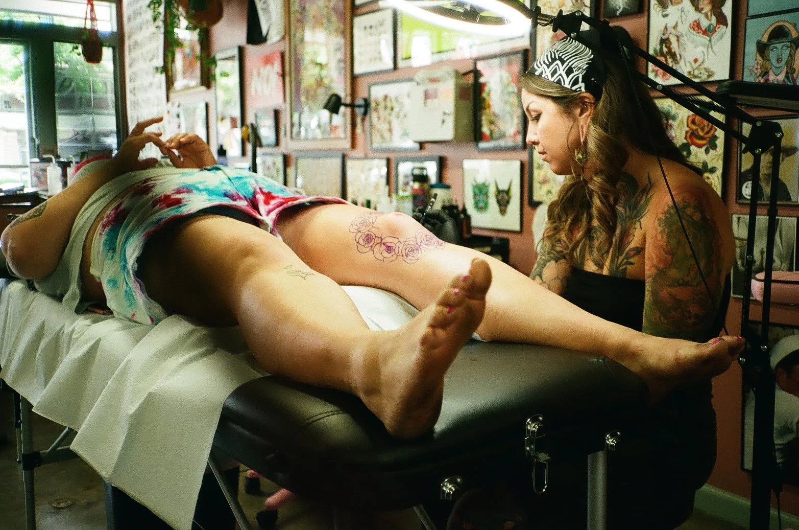 Tattoo artist working on a client's leg in a tattoo studio, with framed artwork on the walls and floral wallpaper in the background.