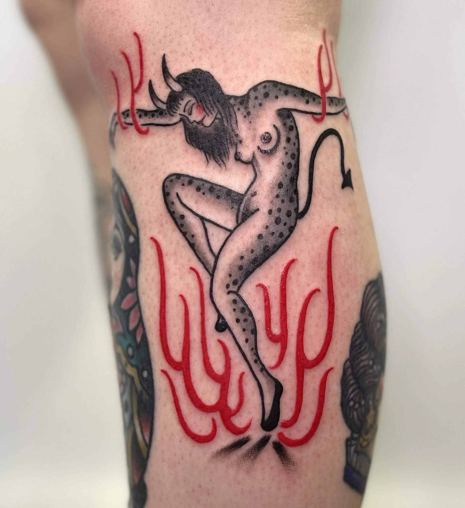 Tattoo of a demon with dancing in the flames, long black hair, and a lizard-like body, surrounded by red flames and some other partial tattoos.