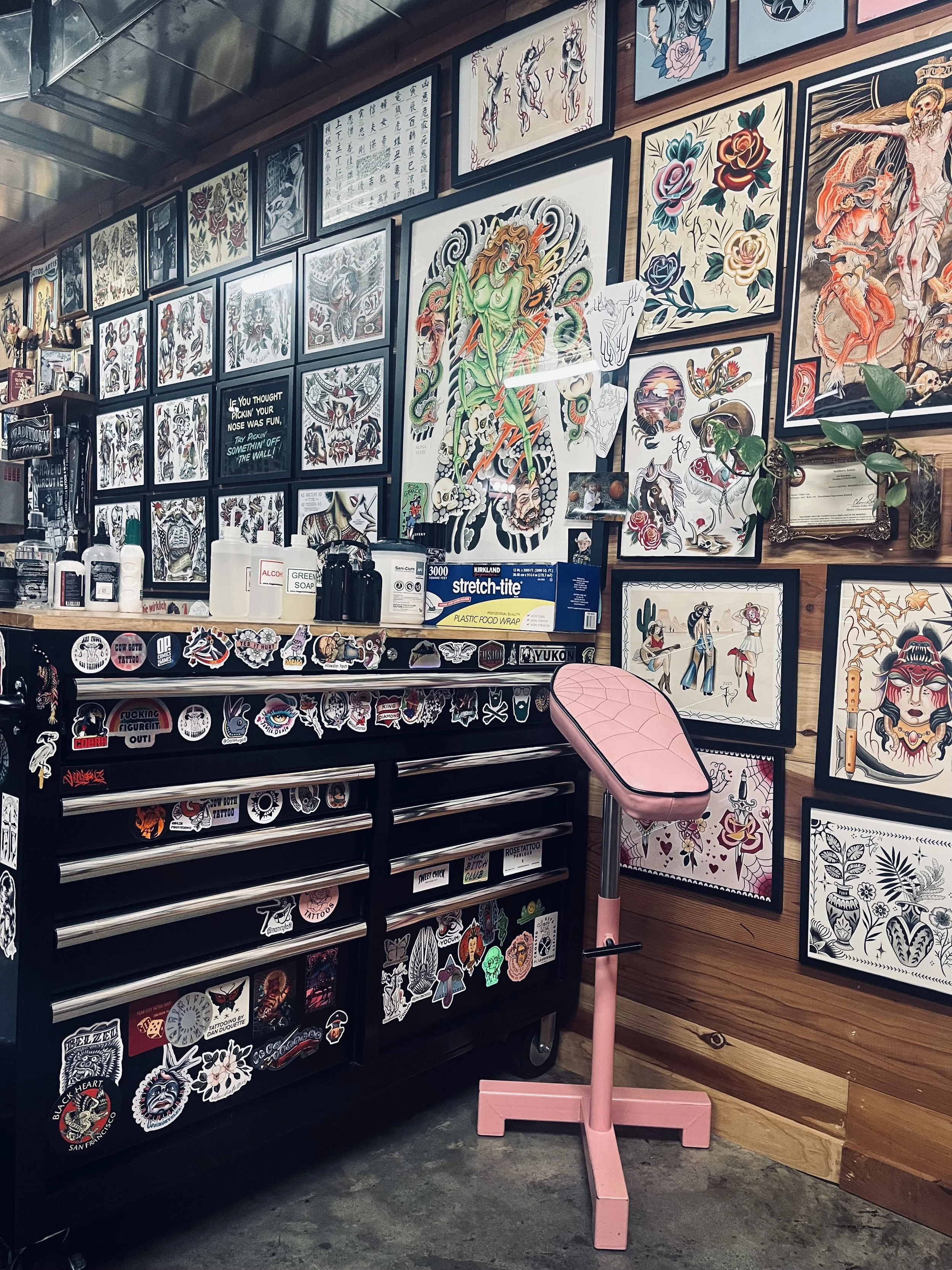 Tattoo shop interior with walls covered in colorful tattoo flash art, drawings, and posters. A pink tattoo chair in front of a black tattoo tool cabinet decorated with various stickers. Various tattoo supplies and equipment on the countertop.