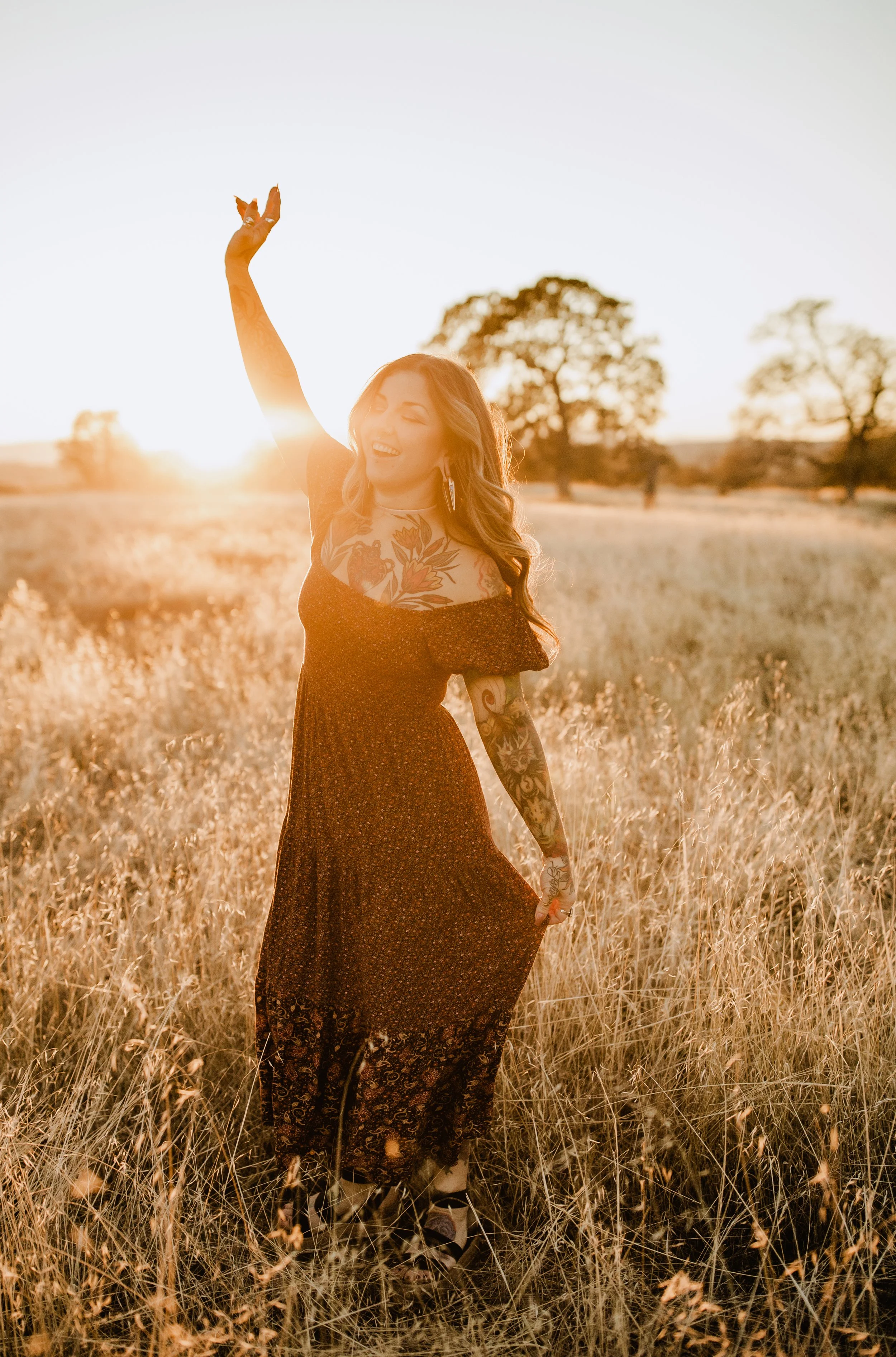 Woman with tattoos dancing in a field at sunset, wearing a long dress.