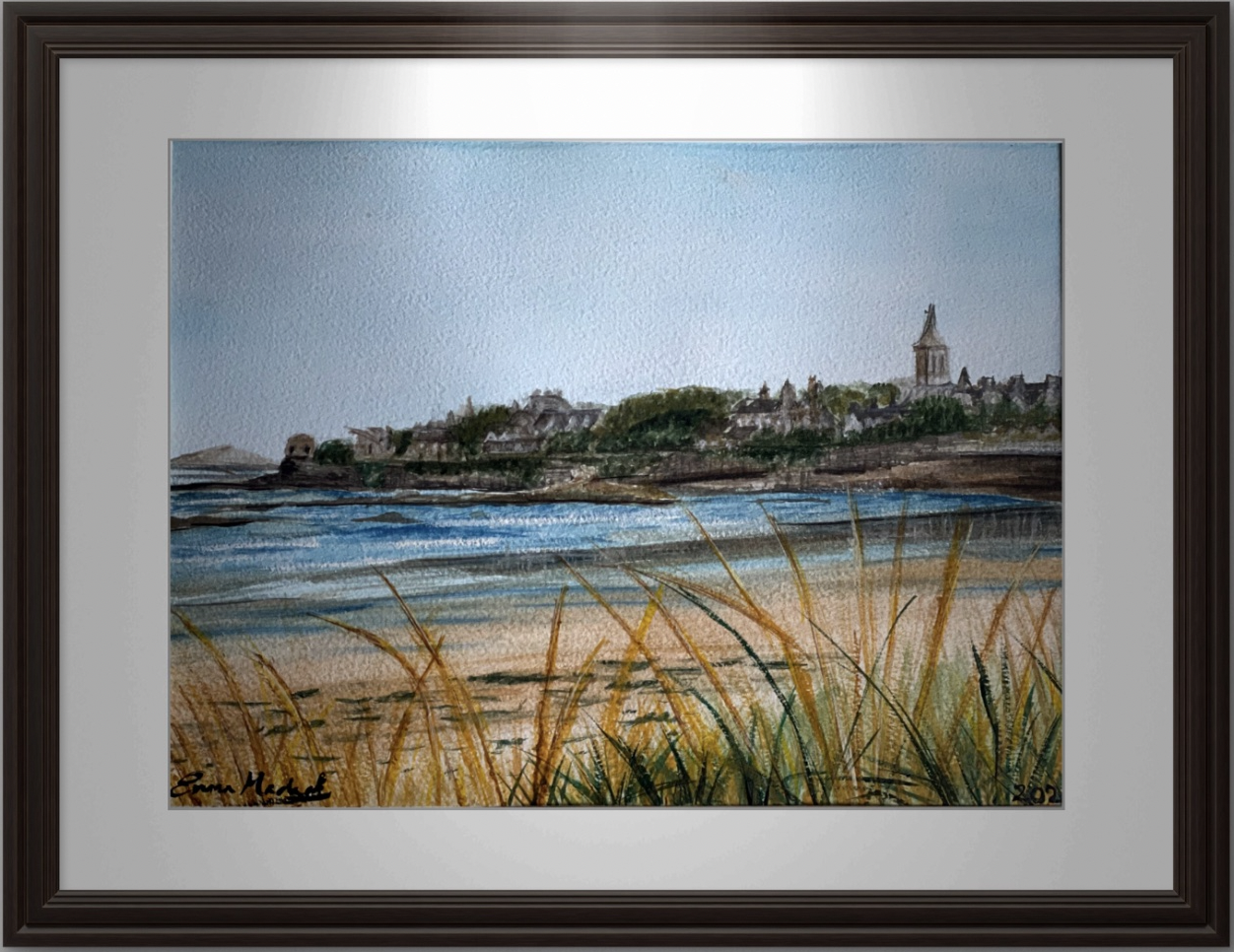 A framed watercolor painting of a coastal town in Scotland viewed from the beach, with grassy dunes in the foreground, houses, and a prominent church steeple in the background.