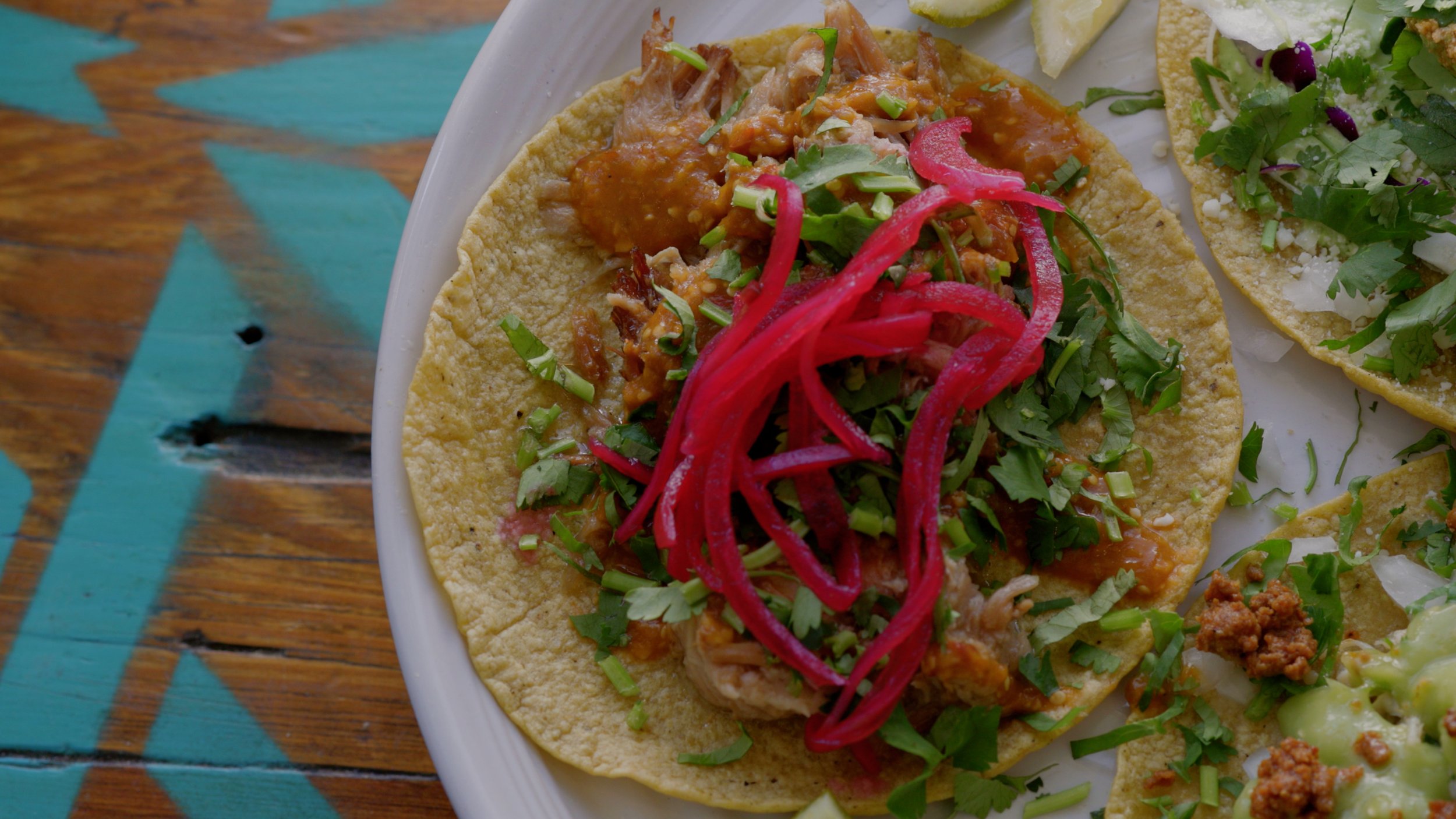 Close-up of a traditional Mexican taco with shredded meat, topped with chopped cilantro, diced onions, and pickled red onions, served on a corn tortilla with lime wedges on the side.