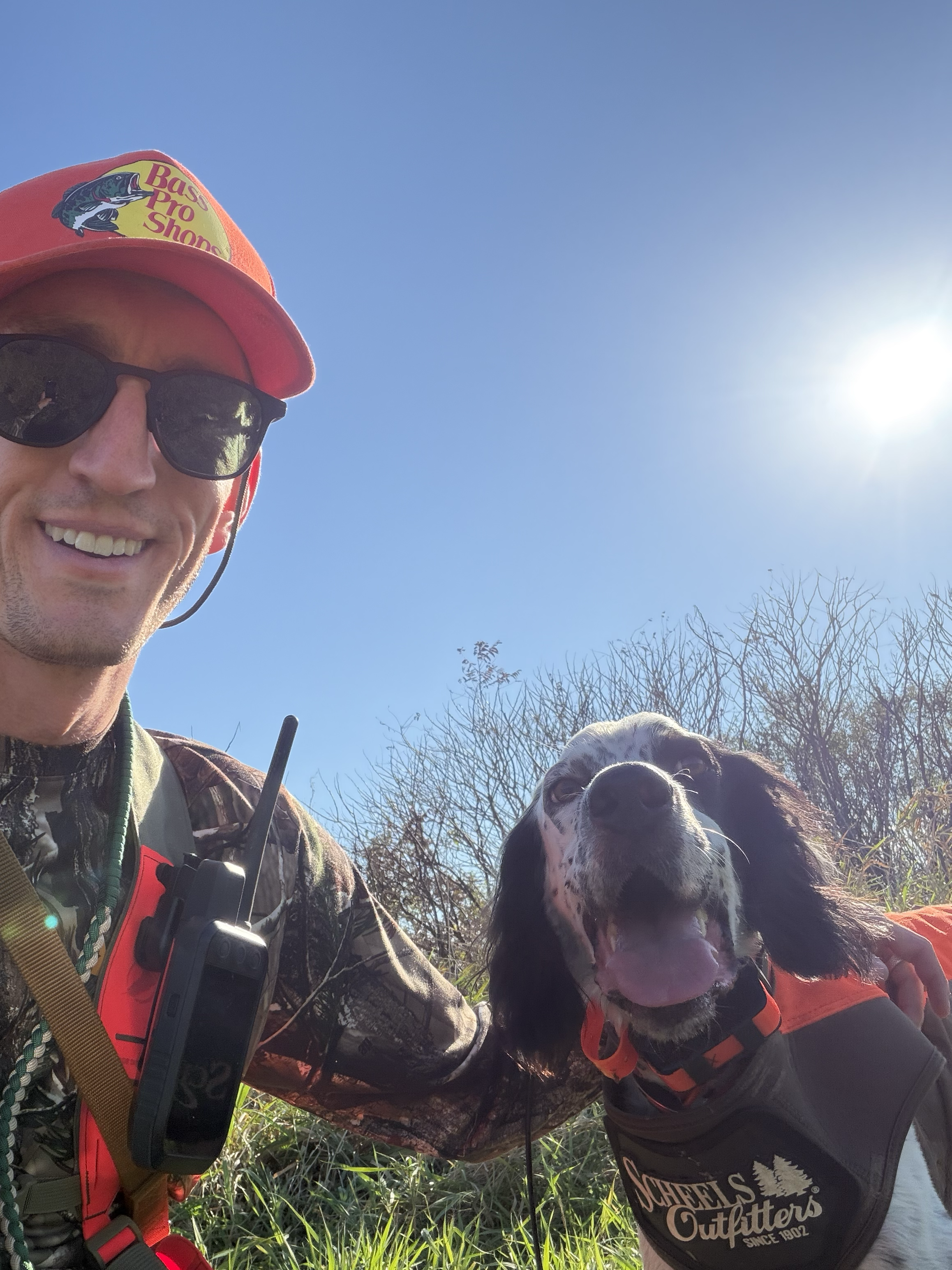 A man wearing a red cap, sunglasses, camouflage shirt, and a walkie-talkie on his chest taking a selfie outdoors with a happy dog wearing a vest that says 'O'Chiels Outfitters'. The sun is shining brightly in a clear blue sky with some dry bushes in the background.