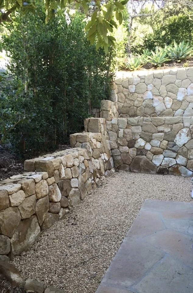 Stone retaining wall with steps, next to a gravel patio area, with green bushes and plants in the background.