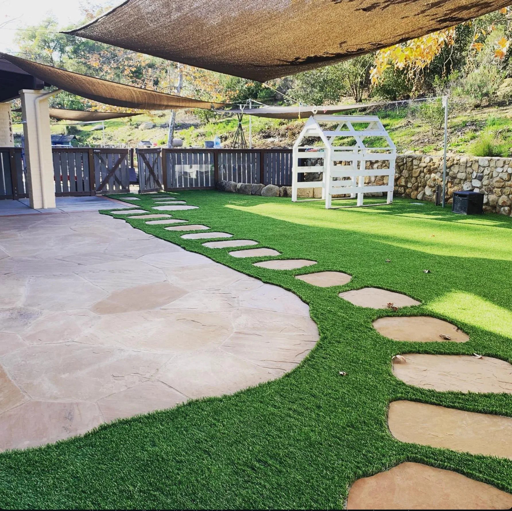 Backyard patio with artificial grass, stepping stones, a white wooden garden structure, and overhead shade sails. There is a stone retaining wall and some outdoor furniture in the background.