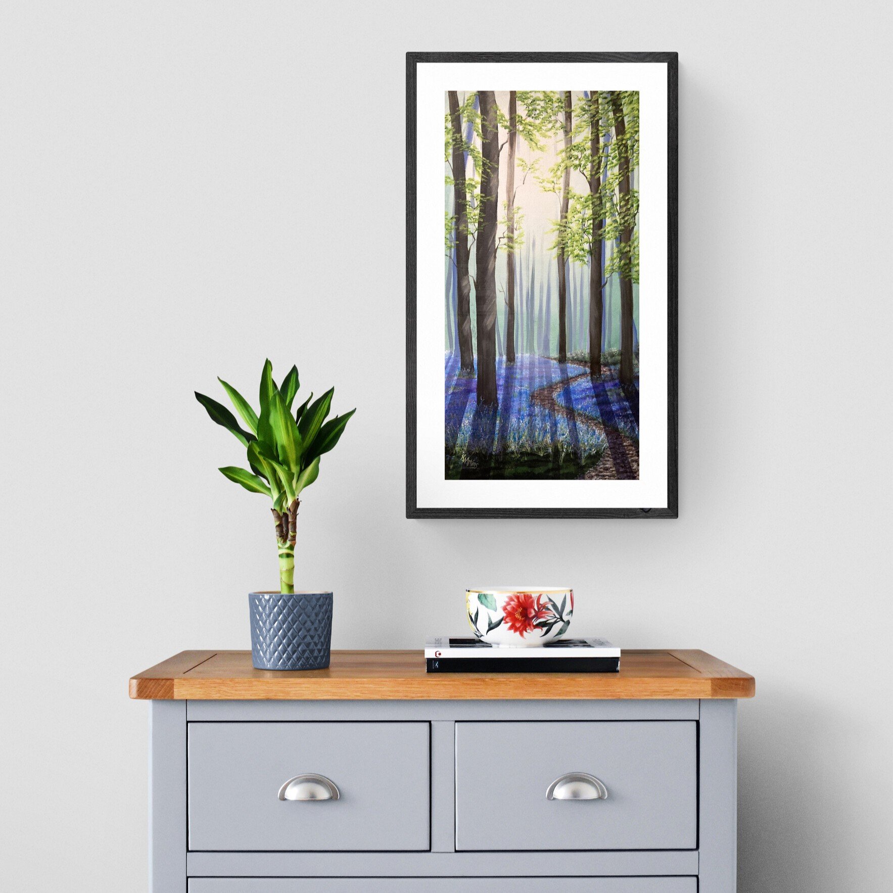 A gray wooden cabinet with two drawers and silver handles, featuring a wooden top surface. On the surface, there's a potted green houseplant in a textured blue pot, and a bowl with a floral design placed on top of a black and white book. Above the cabinet, a framed watercolor painting of a forest scene with trees and sunlight on the ground is hanging on the white wall.
