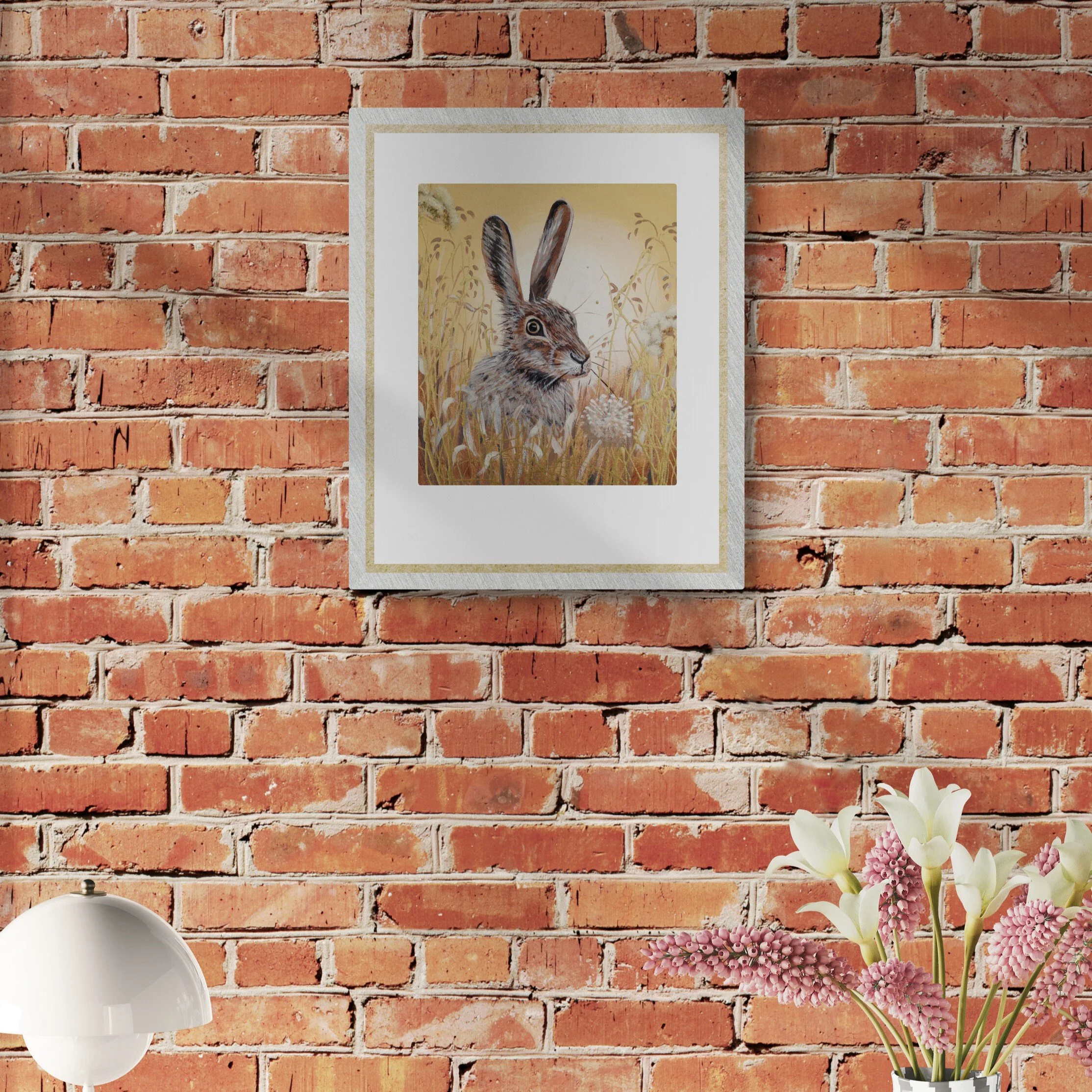 A framed picture of a hare in a field of tall grass and flowers on a red brick wall. There is a white lamp and a vase of pink and white flowers in front of the wall.