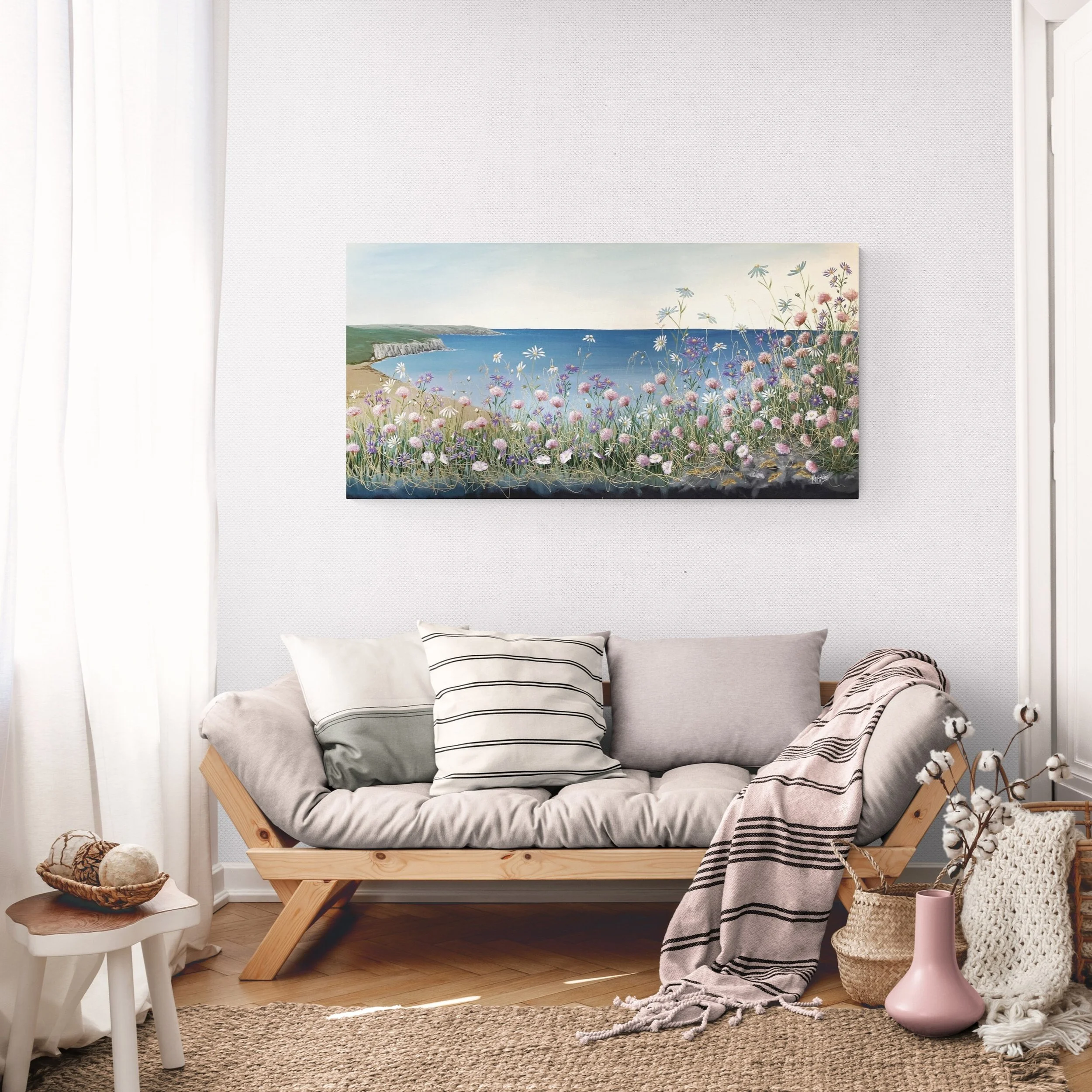 Living room with a wooden sofa, three pillows, a striped blanket, a potted plant, a pink vase, and a basket with yarn, with a large landscape painting of a coastal scene with flowers hanging on the wall.