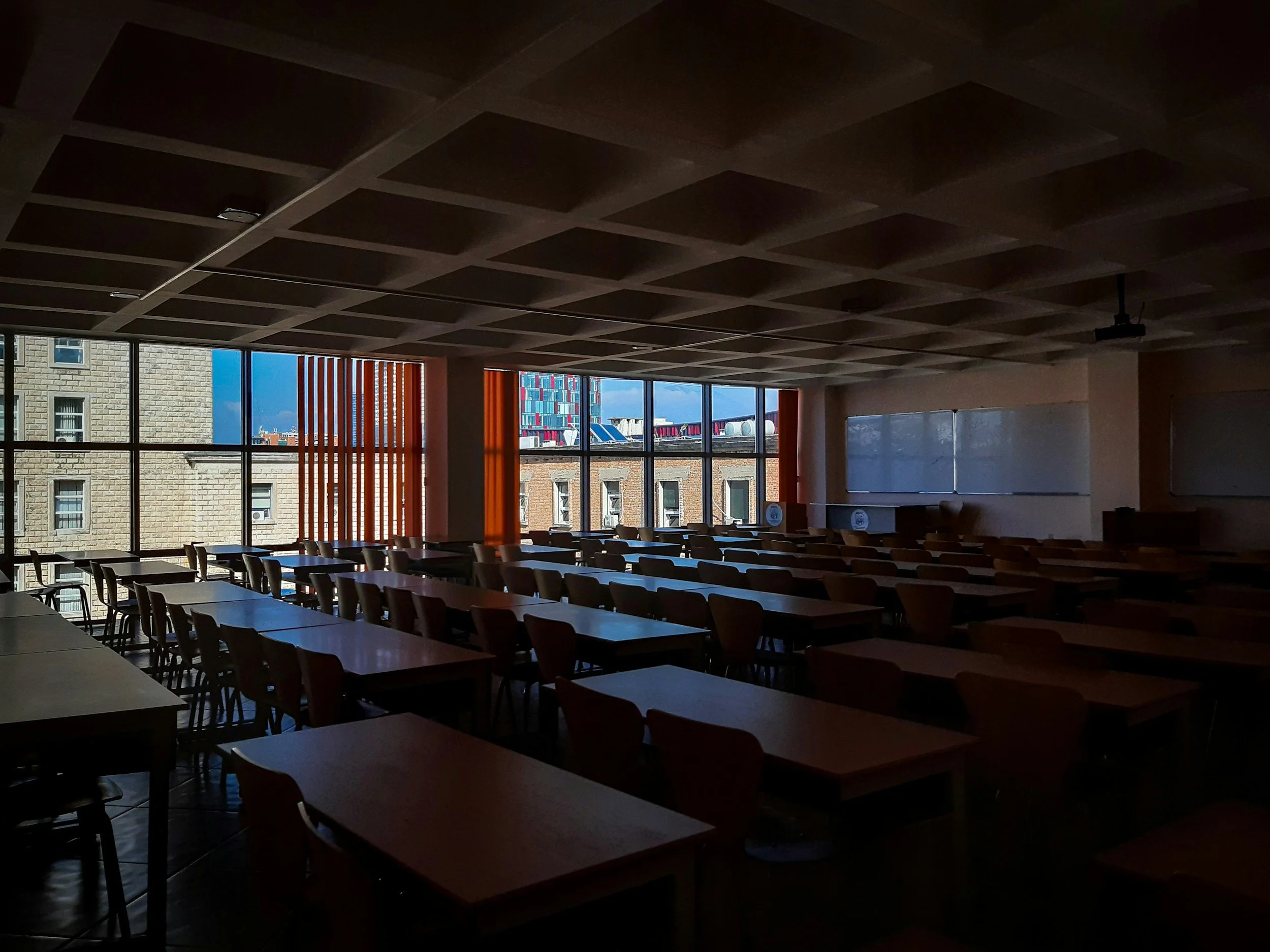 Interior of a classroom or conference room with row of tables and chairs, large windows showing city buildings outside, and a ceiling with grid pattern.