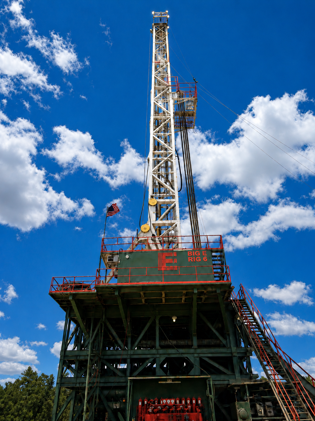 Offshore oil drilling rig with a tall derrick, American flag, and blue sky with white clouds.