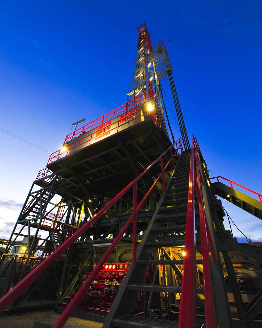 An oil drilling rig with red and yellow safety railings, illuminated lights, and a tall structure against a deep blue evening sky.