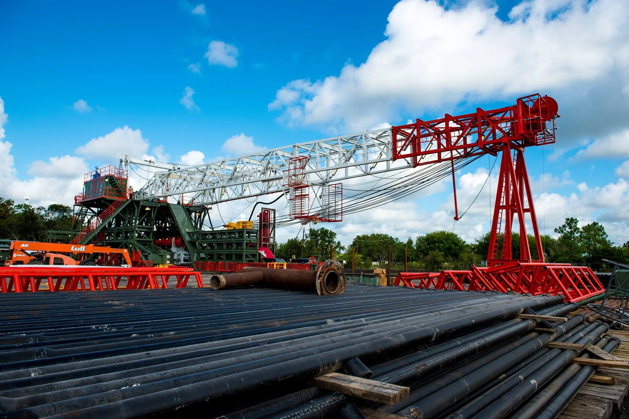 Construction site with black pipes and a large red and white drilling rig under a partly cloudy blue sky.