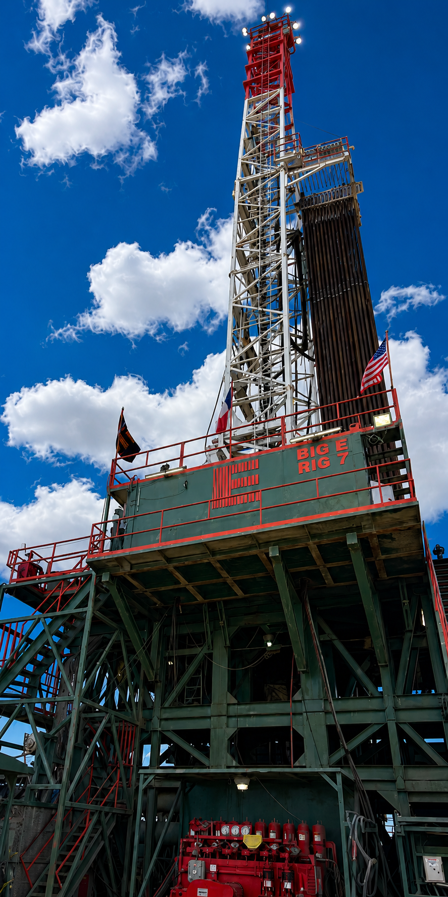 An oil drilling rig named Big E Rig 7, painted in green and red, with a tall metal tower, American flag, and equipment underneath, set against a bright blue sky with clouds.