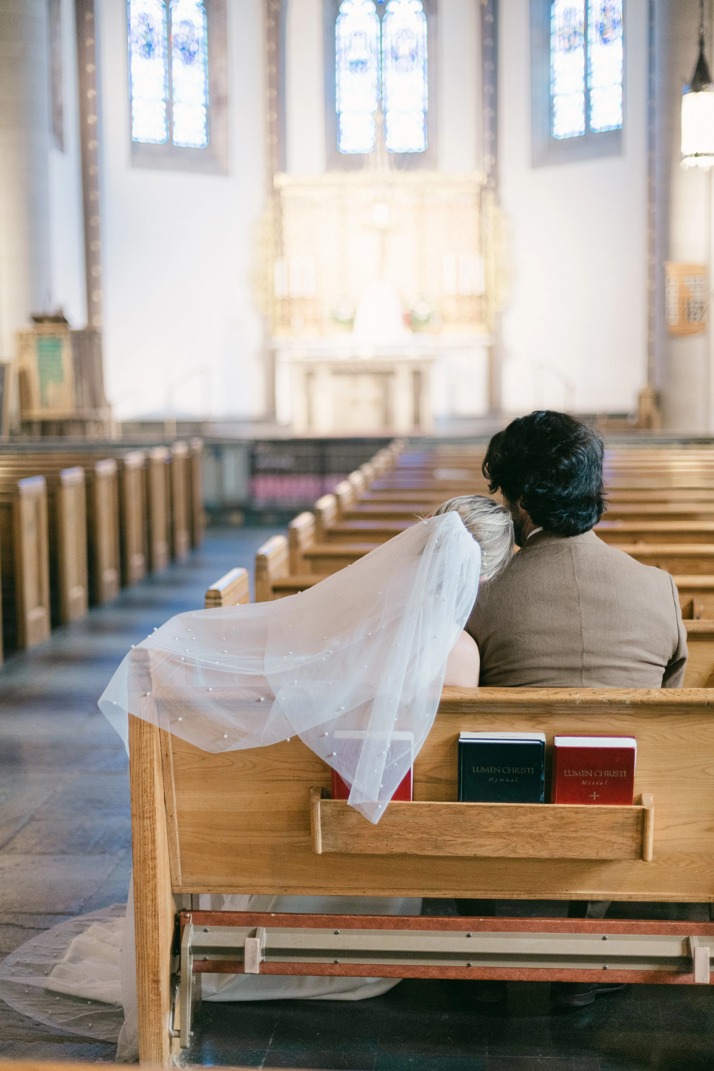 A bride and groom sitting together in a church pew, with the bride leaning her head on the groom's shoulder. The bride has a veil, and the church interior features stained glass windows and an altar in the background.