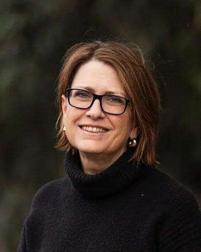 Portrait of a woman wearing glasses and a black turtleneck sweater, smiling outdoors with a blurred background.