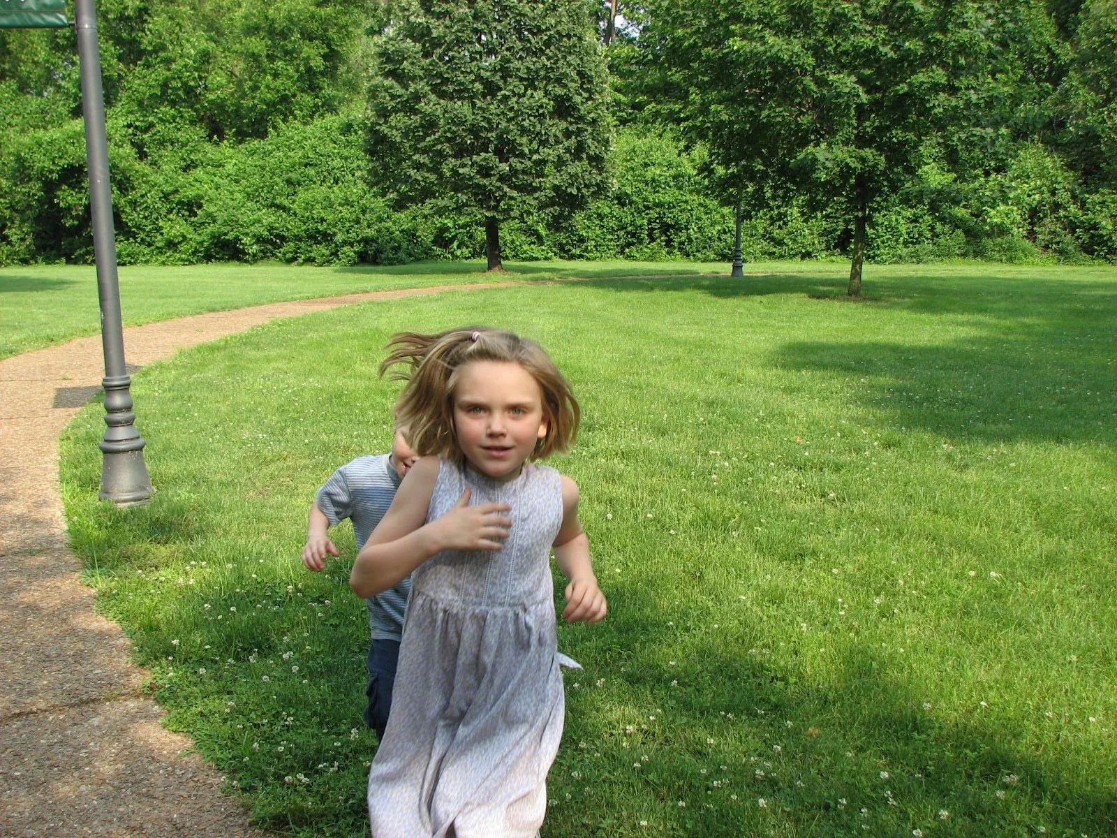 Two children, a girl and a boy, running in a park on a sunny day, with trees and a lamp post in the background.