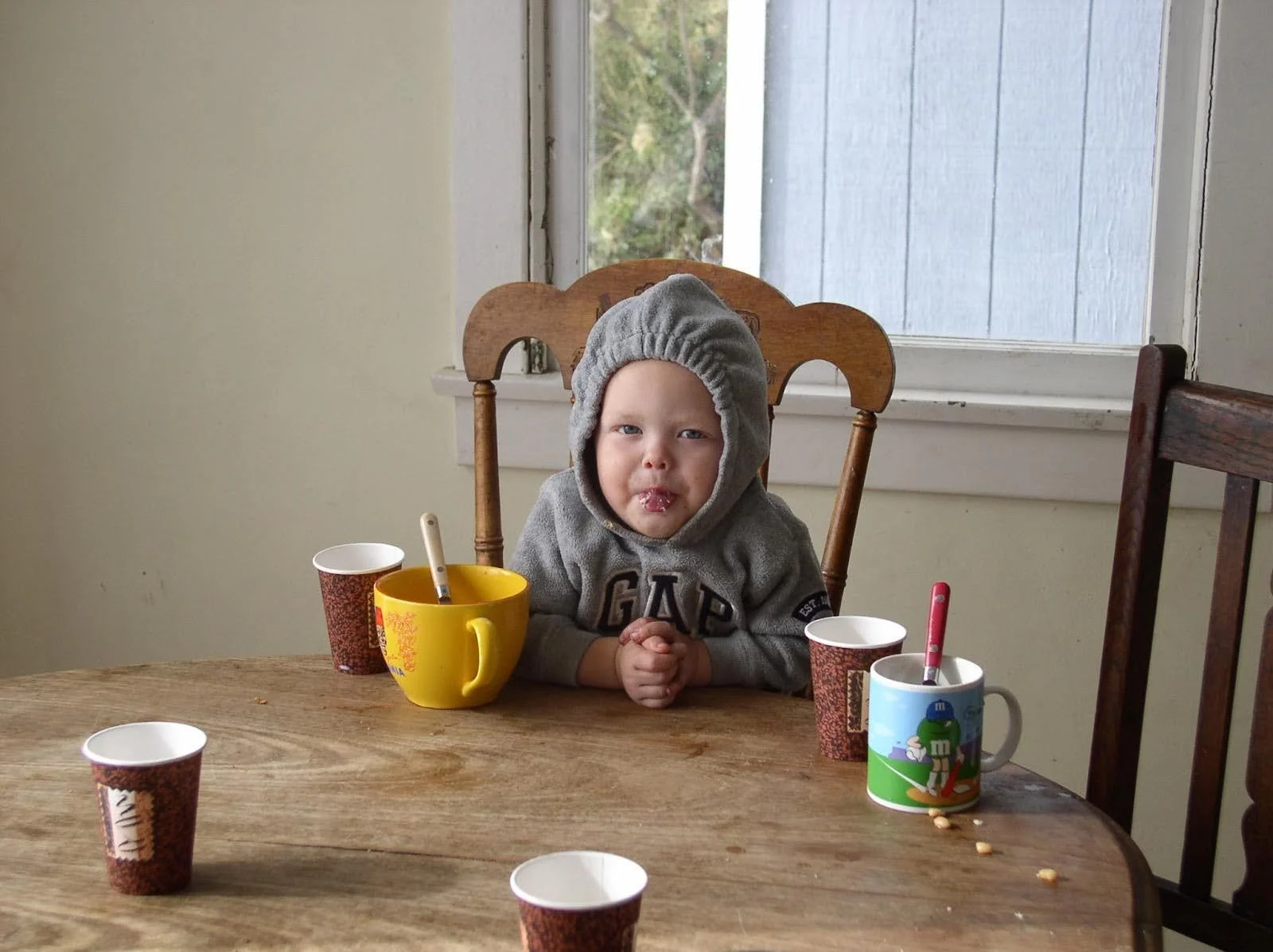 A young child sitting at a wooden table in a high chair, wearing a gray hoodie with the hood up, and sticking out their tongue with some food inside. There are four paper cups and a yellow mug on the table, along with some snacks.