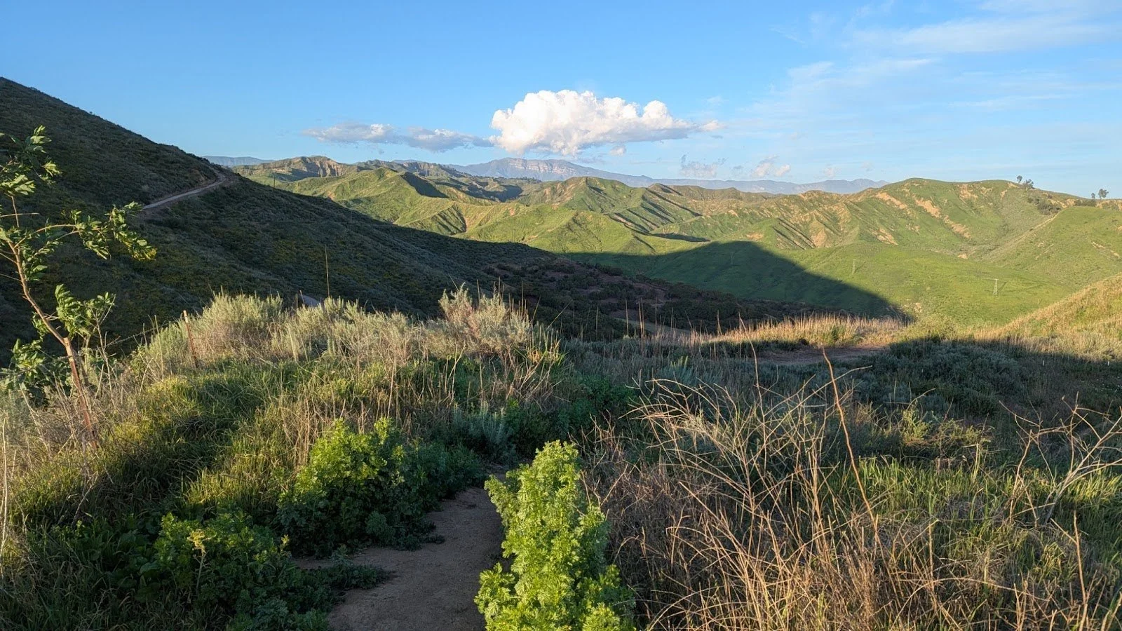 View of green rolling hills under a clear blue sky with scattered white clouds, some shadows cast on the landscape.