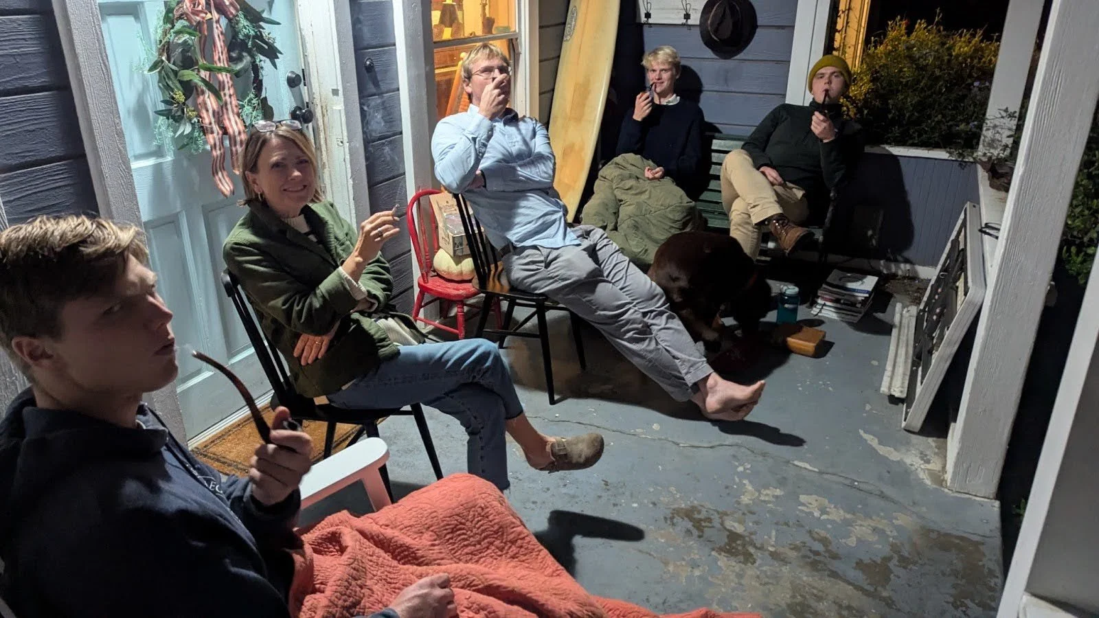 A group of seven people sitting on chairs and a bench on a porch at night, some smoking and talking, with a dog lying on the floor, and various objects like books and wood around.