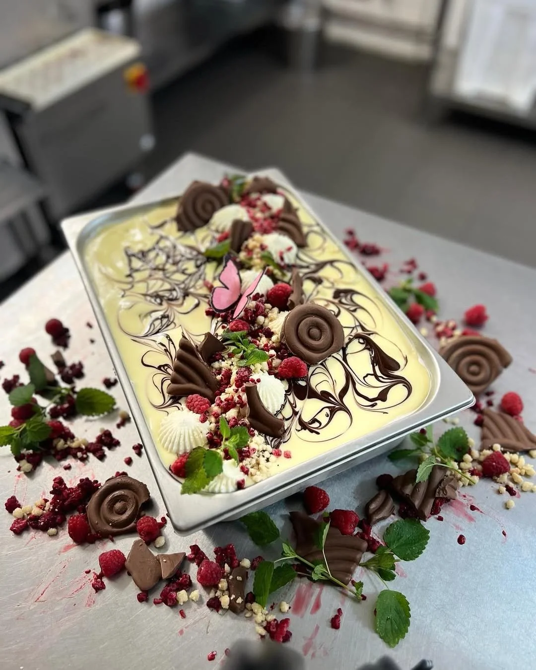 A decorated ice cream or gelato dessert topped with chocolate swirls, white and pink flowers, red berries, and green mint leaves, with additional toppings scattered around on a metal surface.