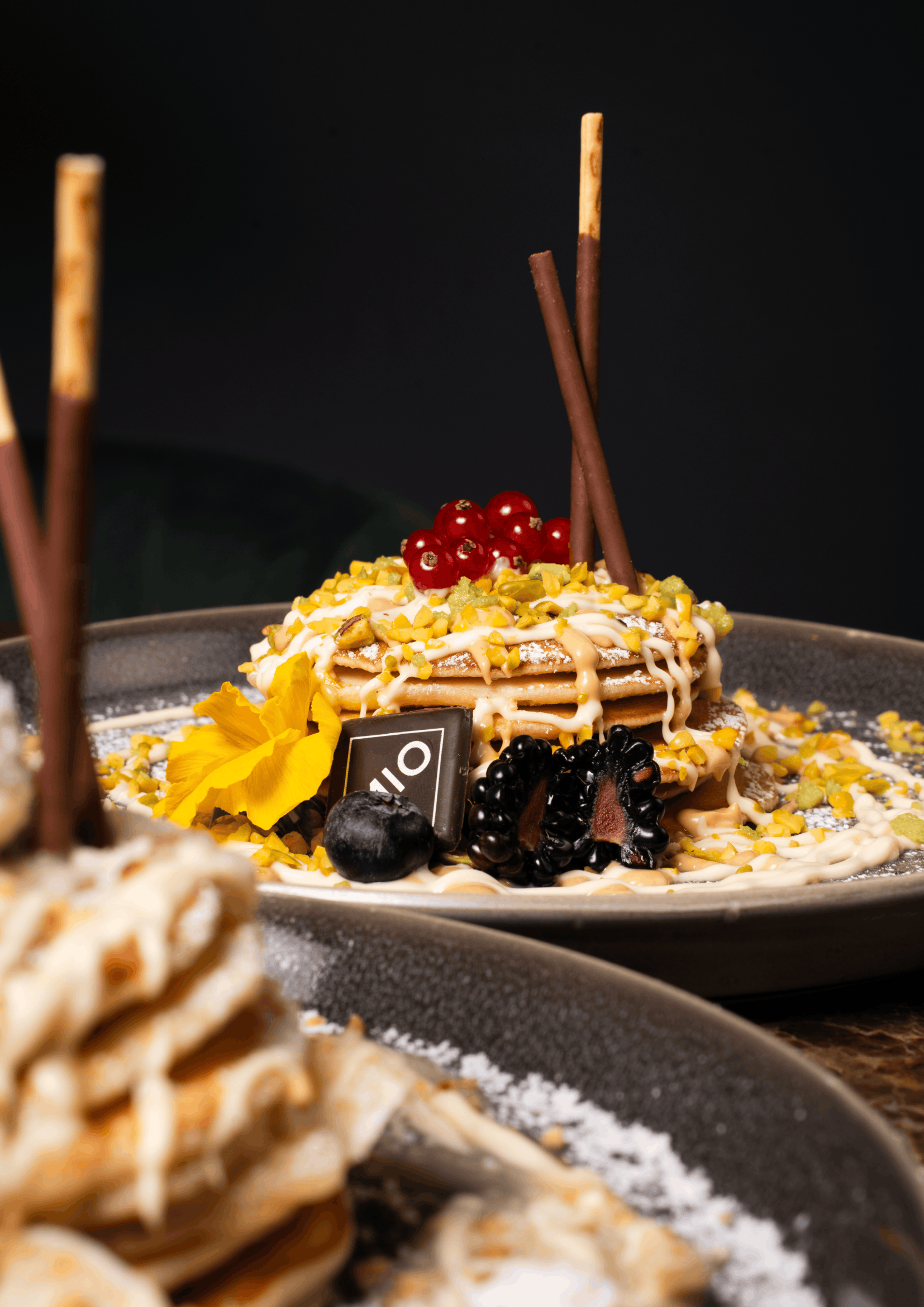 Stack of pancakes topped with berries, chopped pistachios, garnished with flower, chocolate pieces, and drizzled with sauce, served on a black plate.