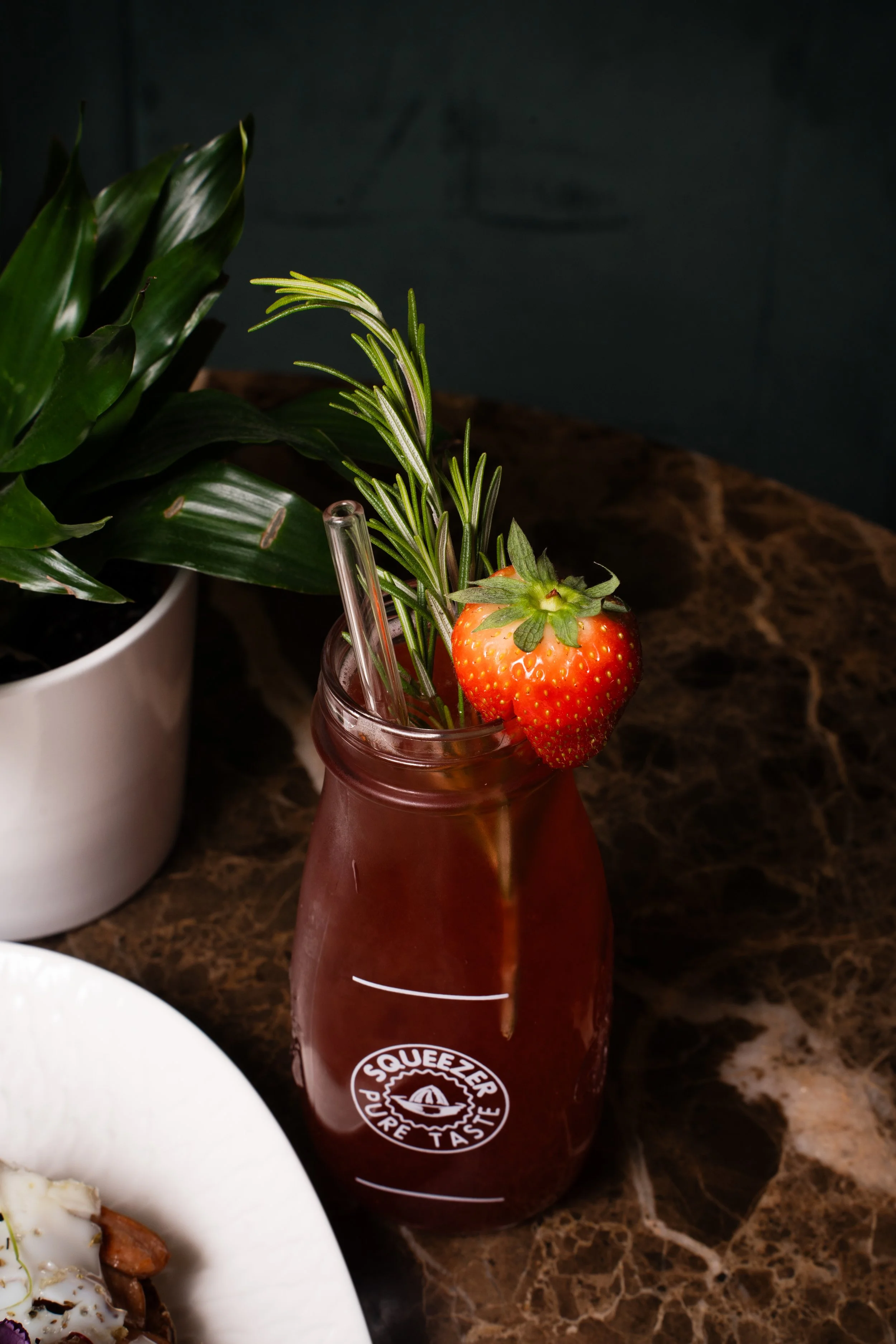 A glass bottle with the logo 'Squeeze Pure Taste' filled with red liquid, garnished with a strawberry and sprigs of rosemary and thyme, placed on a dark marble surface.