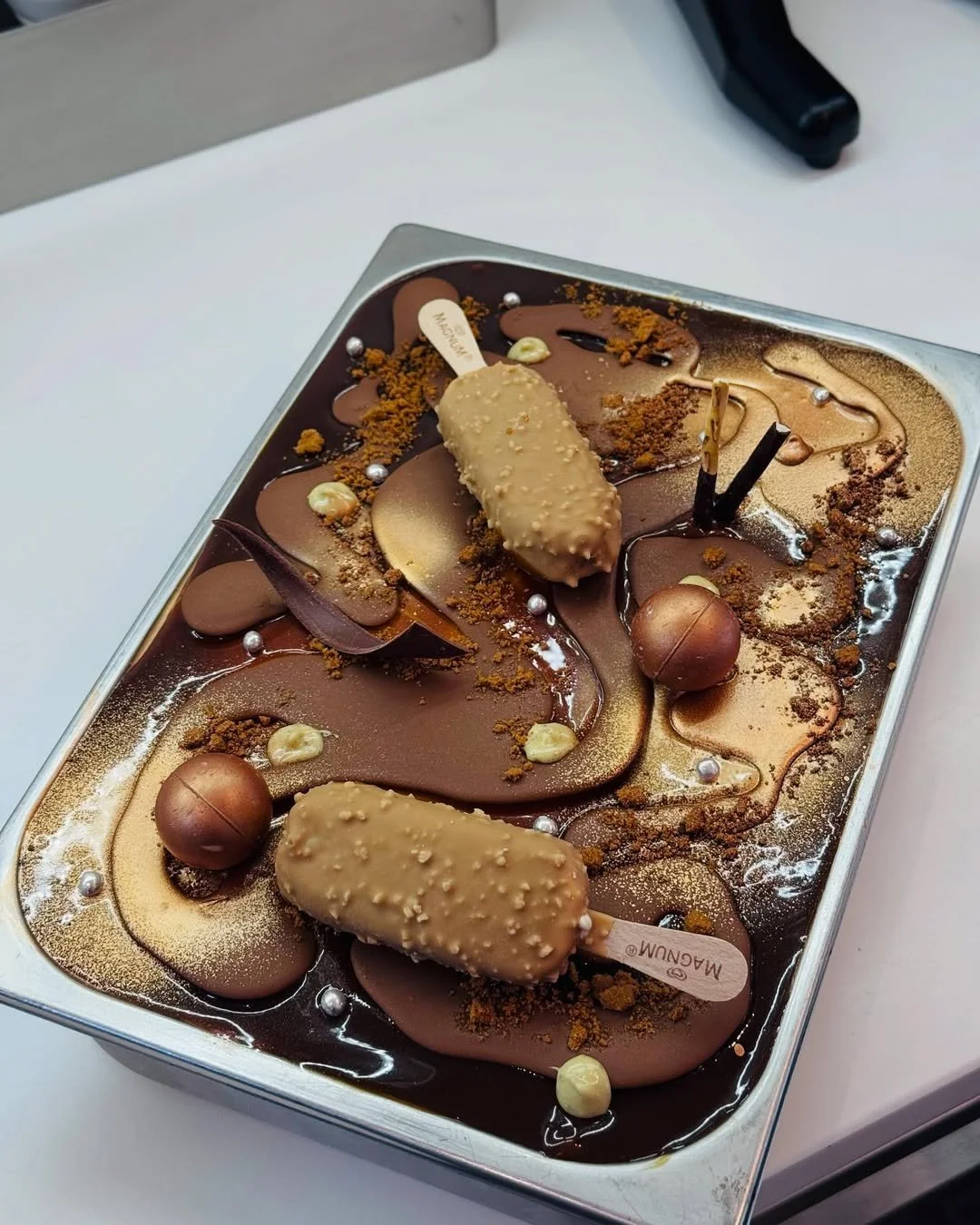 A tray of decorated chocolate ice cream with caramel and chocolate swirls, white and brown chocolate balls, and various crumbs and sprinkles.
