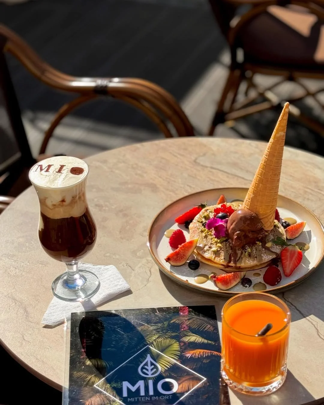 A plate of pancakes topped with ice cream, fresh strawberries, blueberries, and edible flowers, with an upside-down ice cream cone on top. There are two drinks on the table: a dark beverage in a glass with whipped cream and a coffee stirrer, and an o