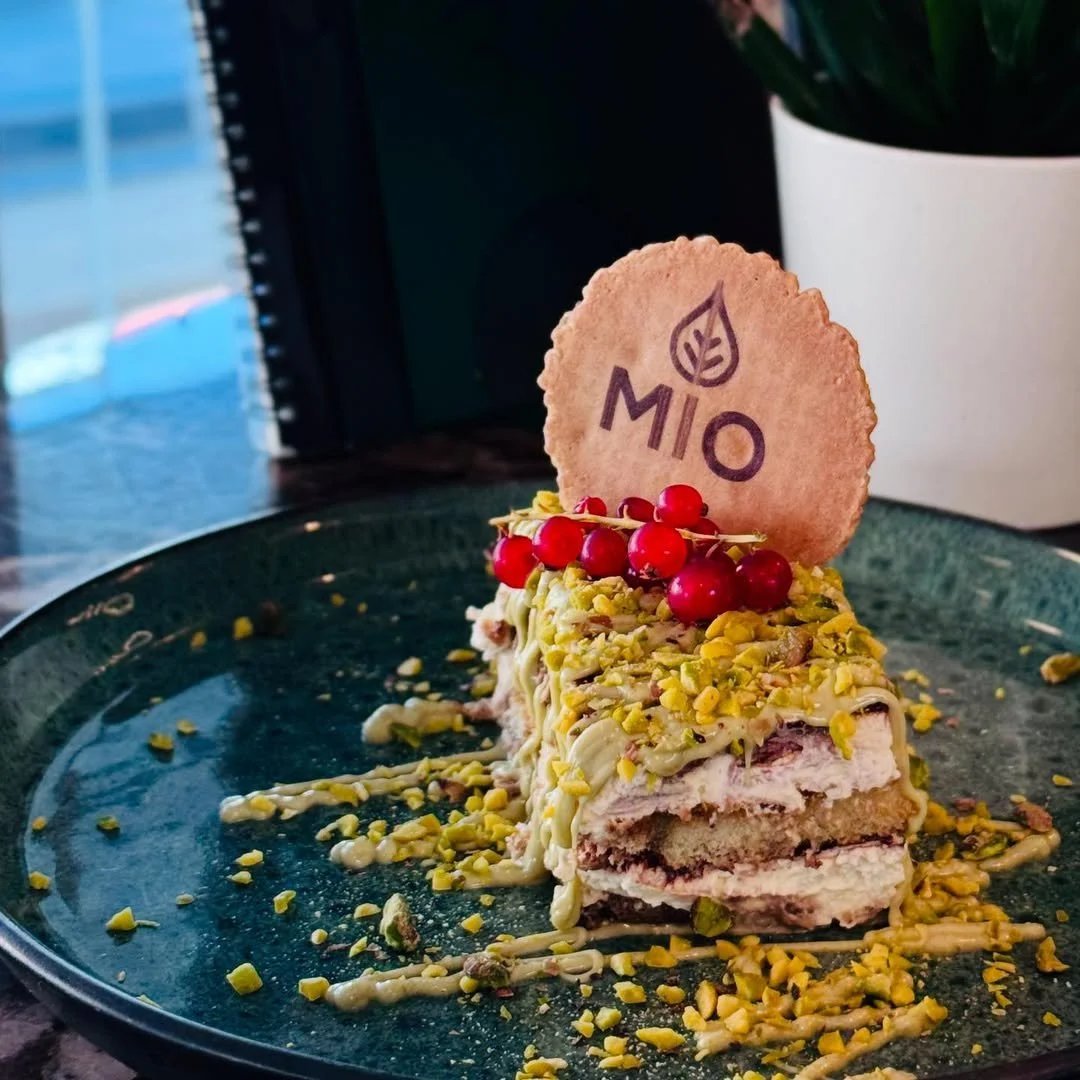 A layered dessert on a black plate, garnished with chopped pistachios, white chocolate drizzle, red berries, and a round cookie with the word 'MIO' and a leaf logo, set against a background with a plant and a window.