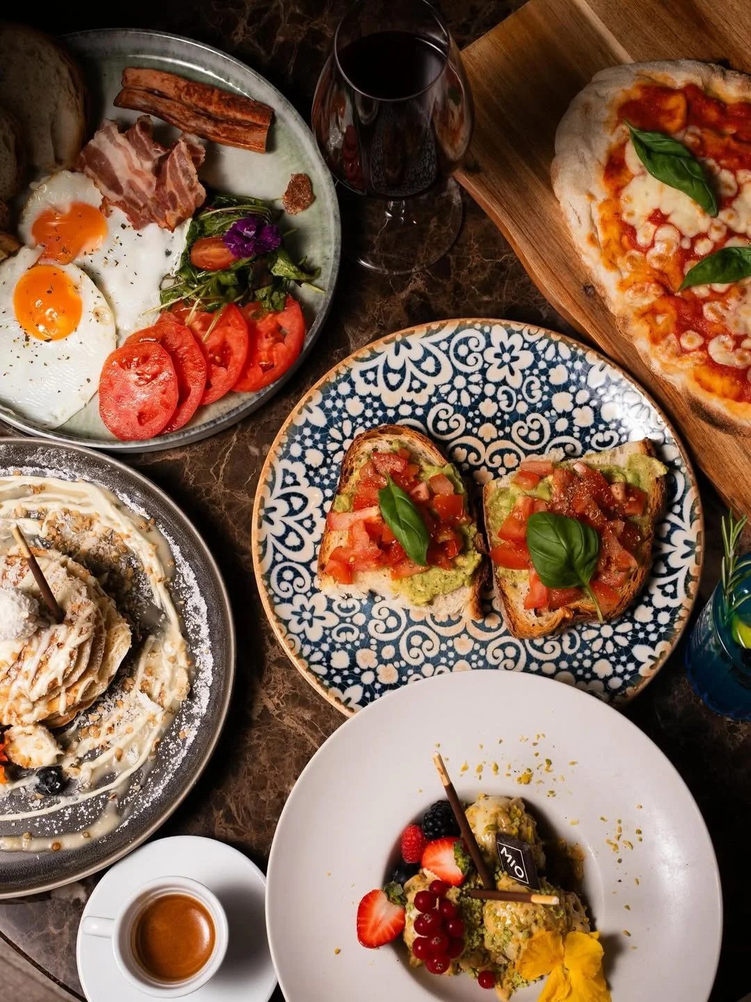 A variety of dishes on a table, including a pizza with basil, avocado toast topped with tomatoes and basil, pasta with white sauce and black olives, a breakfast plate with eggs, bacon, tomatoes, and greens, and a dessert with strawberries, blackberri