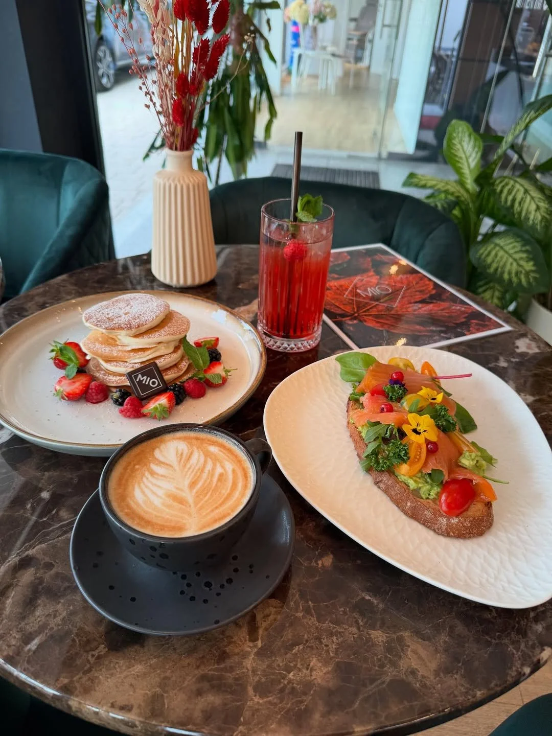 A marble table with a cup of latte, a plate of pancakes with berries, a glass of red fruit drink, and an open-faced sandwich with vegetables, in a cozy cafe setting with plants and a vase of dried flowers.