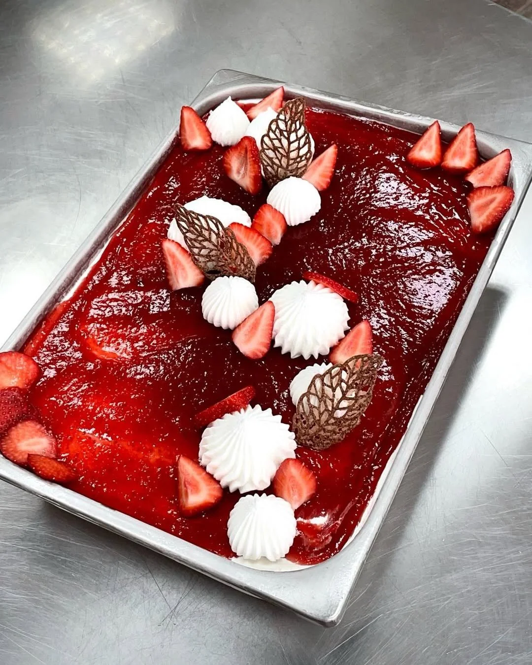 A rectangular tray of strawberry dessert decorated with sliced strawberries, whipped cream, and decorative chocolate leaves.
