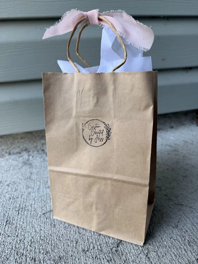 A brown paper shopping bag with twisted paper handles, decorated with light pink fabric tied in a bow at the top, sitting on a textured surface against a light-colored wooden wall. The bag has a circular black logo that reads 'Created by Jissi' with a decorative leaf element.