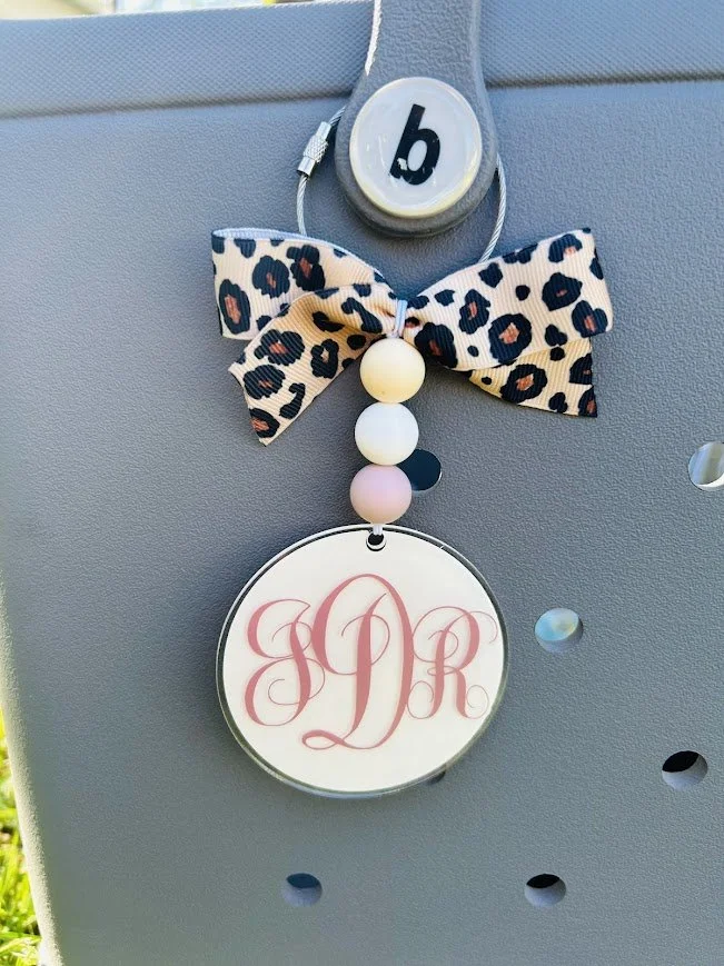 Decorative monogrammed bag tag with pink initials 'JRD' hanging from a string, attached to a Bogg Bag with a leopard print bow and pastel beads.