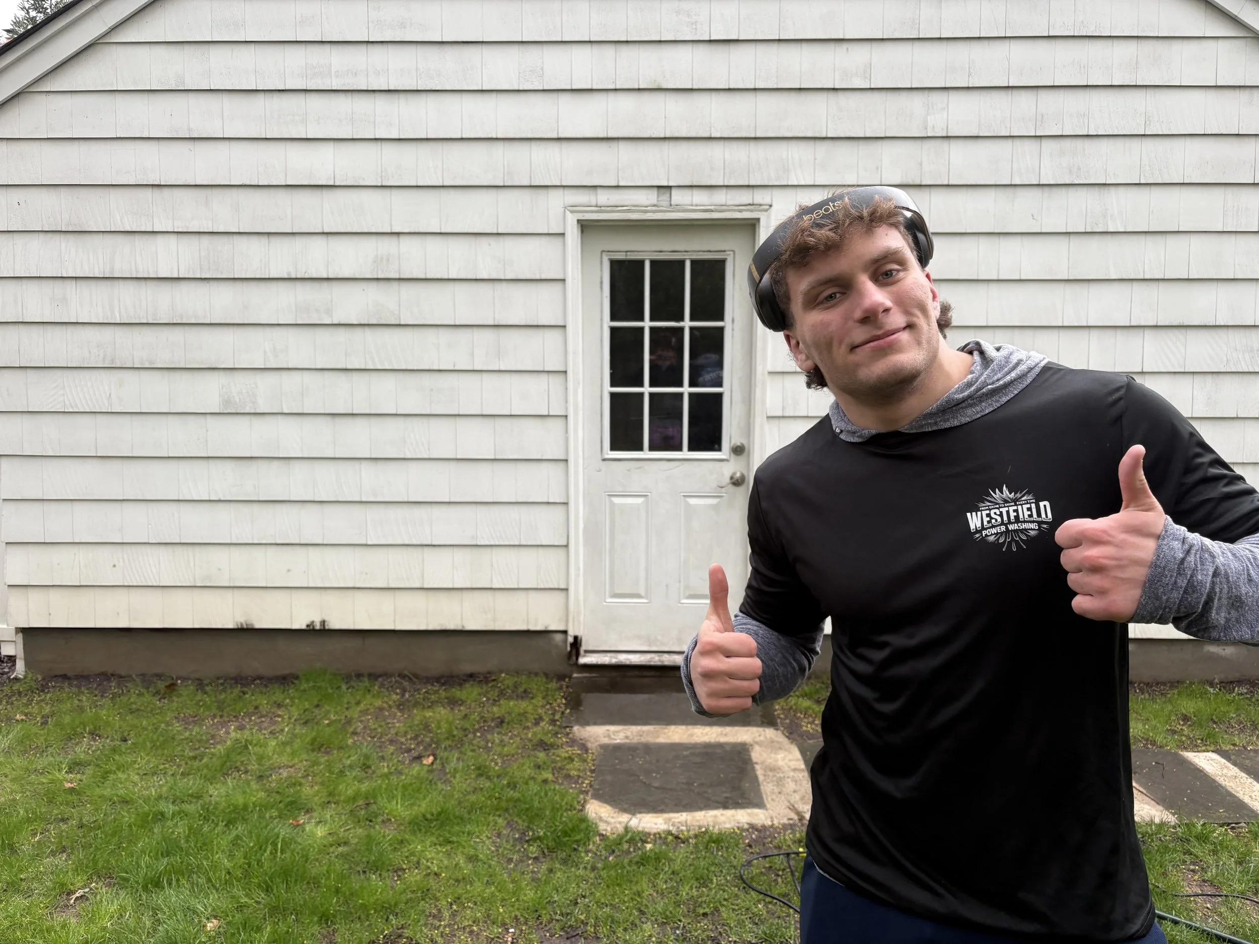A young man wearing headphones and a black T-shirt with Westfield Power Washing logo, giving thumbs up and smiling outdoors in front of a white house with a door, on a grassy area.
