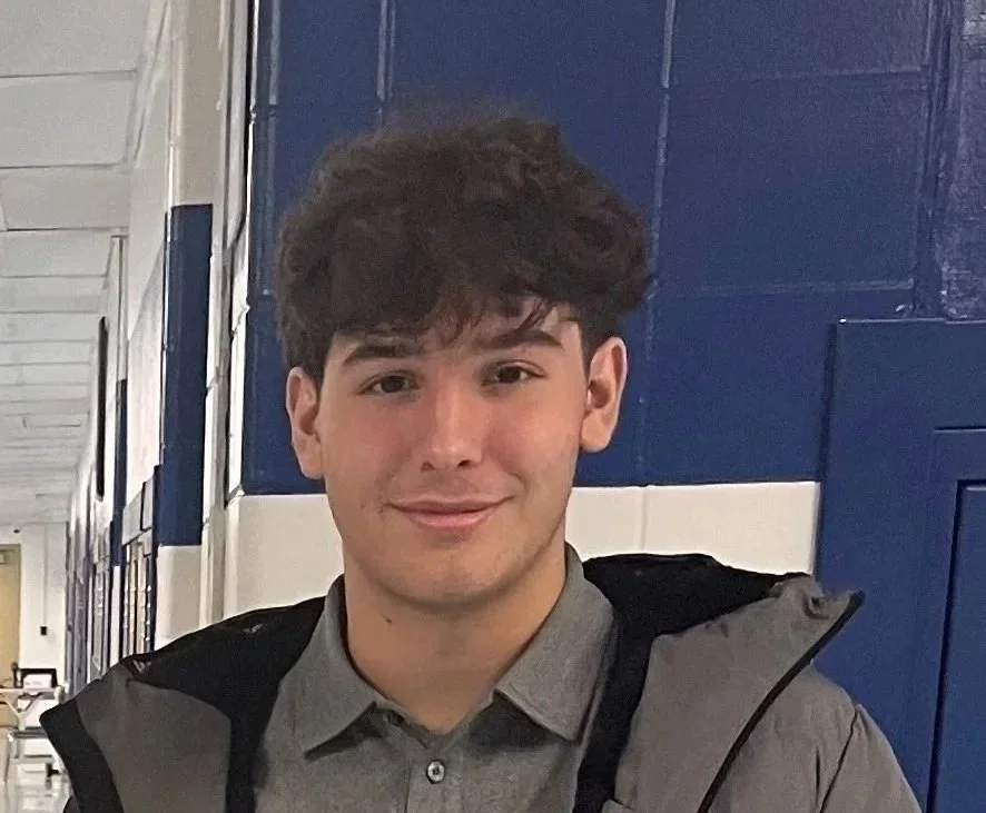 A young man with dark, curly hair smiling in front of a blue and white wall.