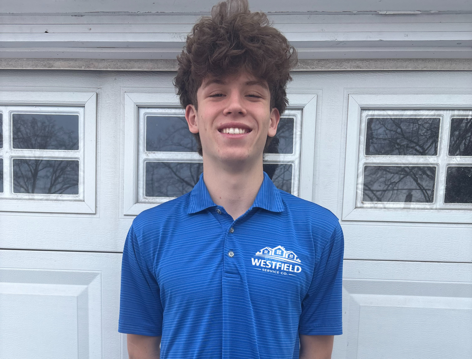 Young man with curly brown hair smiling, wearing a blue polo shirt with 'Westfield Service Co.' logo, standing in front of a white garage door.