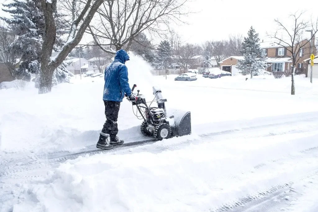 A person in a blue jacket using a snowblower to clear snow from a residential sidewalk during winter.