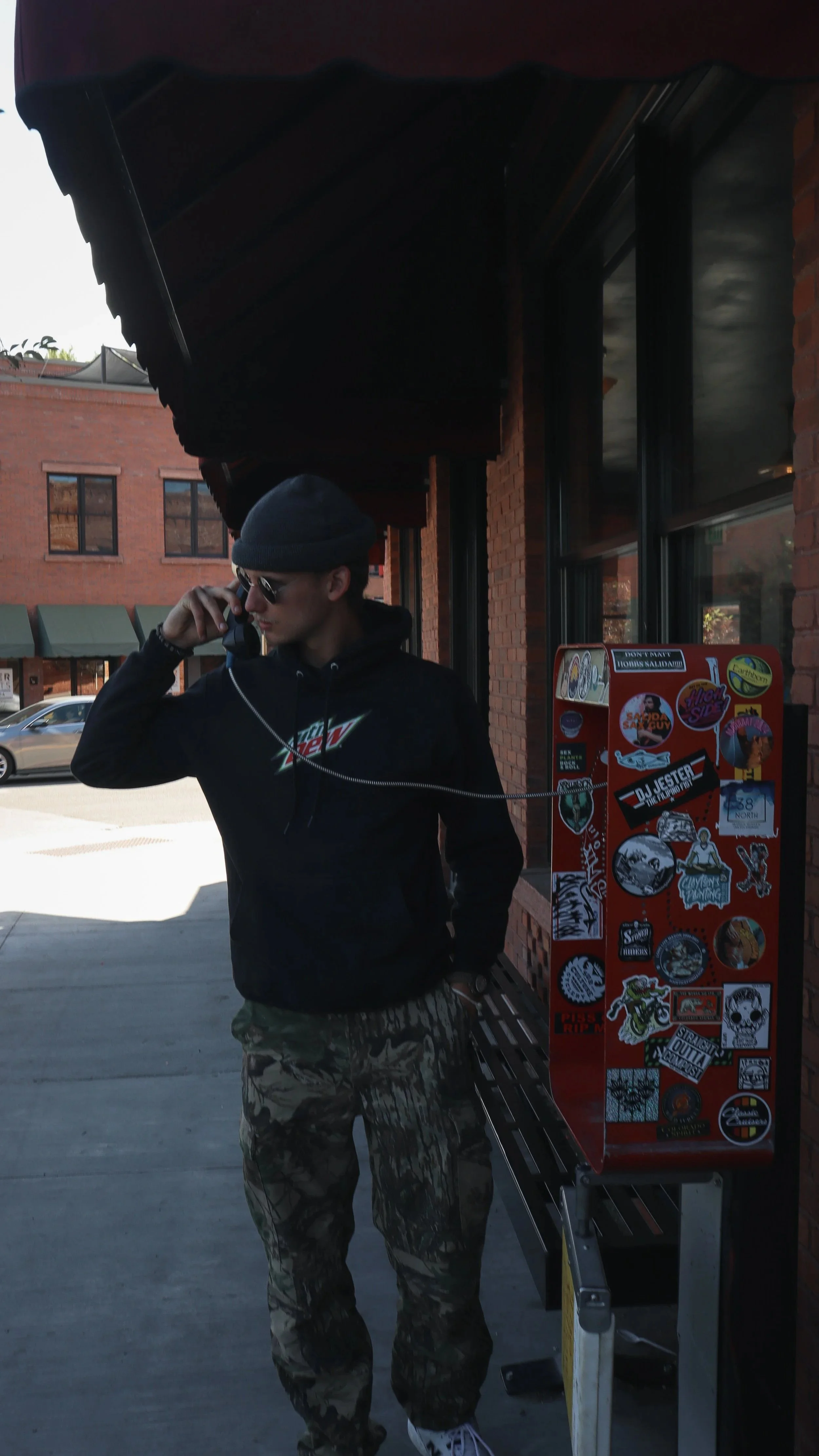 A young man wearing camouflage pants, a black hoodie with a mountain logo, black beanie, and sunglasses is talking on a payphone outside a brick building with a sticker-covered red box beside him.