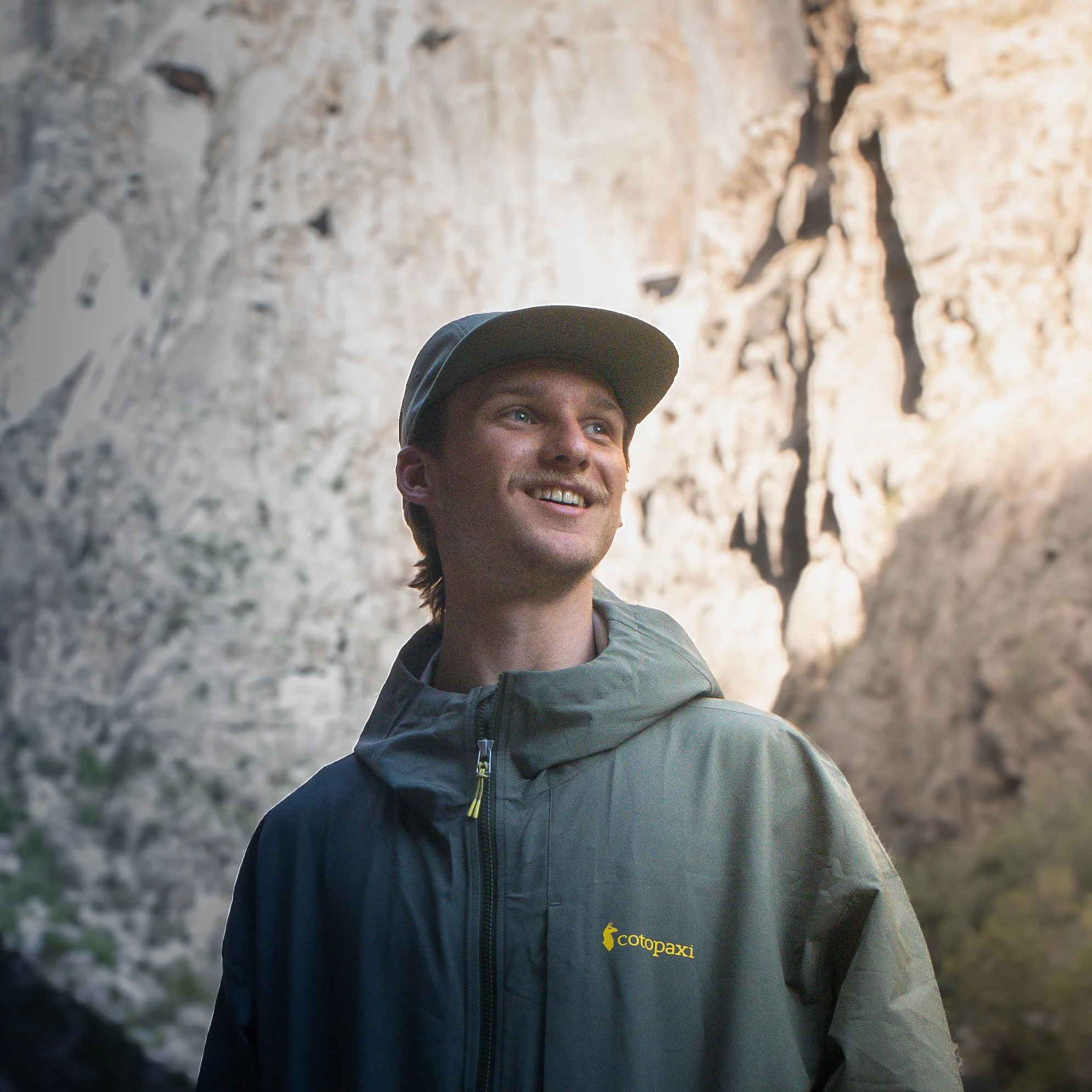 A smiling man in outdoor gear and a baseball cap standing in front of a rocky canyon wall with a shadow of a climber on the cliff face behind him.