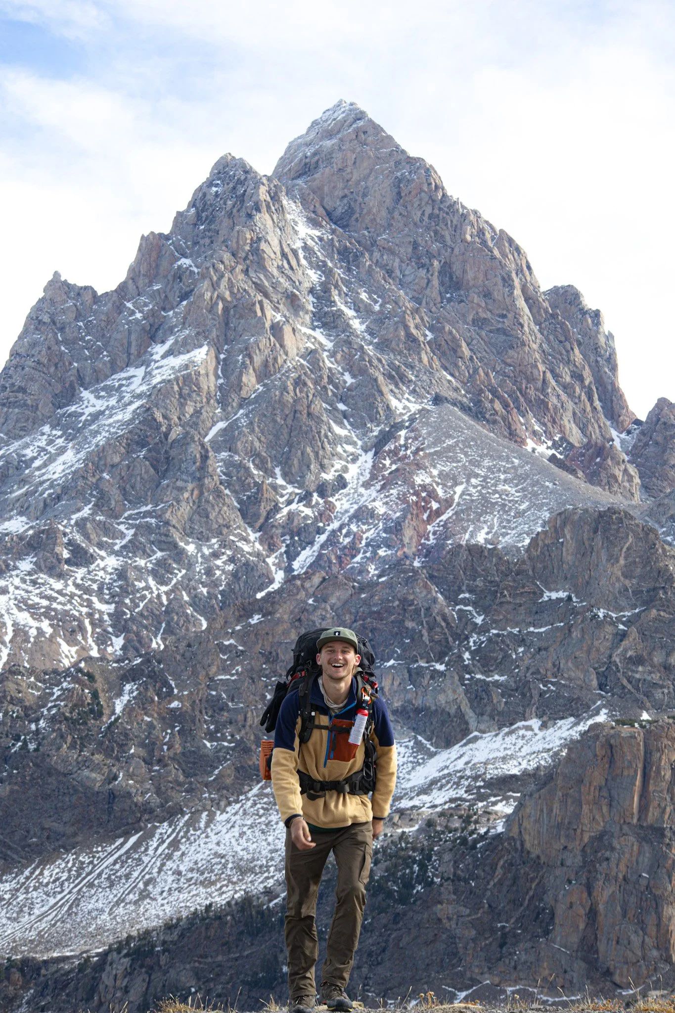 A man hiking in front of a snow-covered mountain with rocky cliffs, wearing a beige jacket, brown pants, and carrying a large backpack.