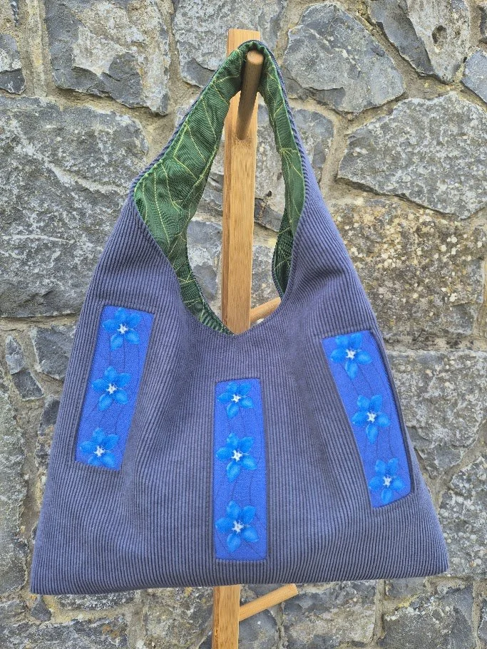 Gray fabric tote bag with blue floral embroidery hanging on a wooden hook against a stone wall.
