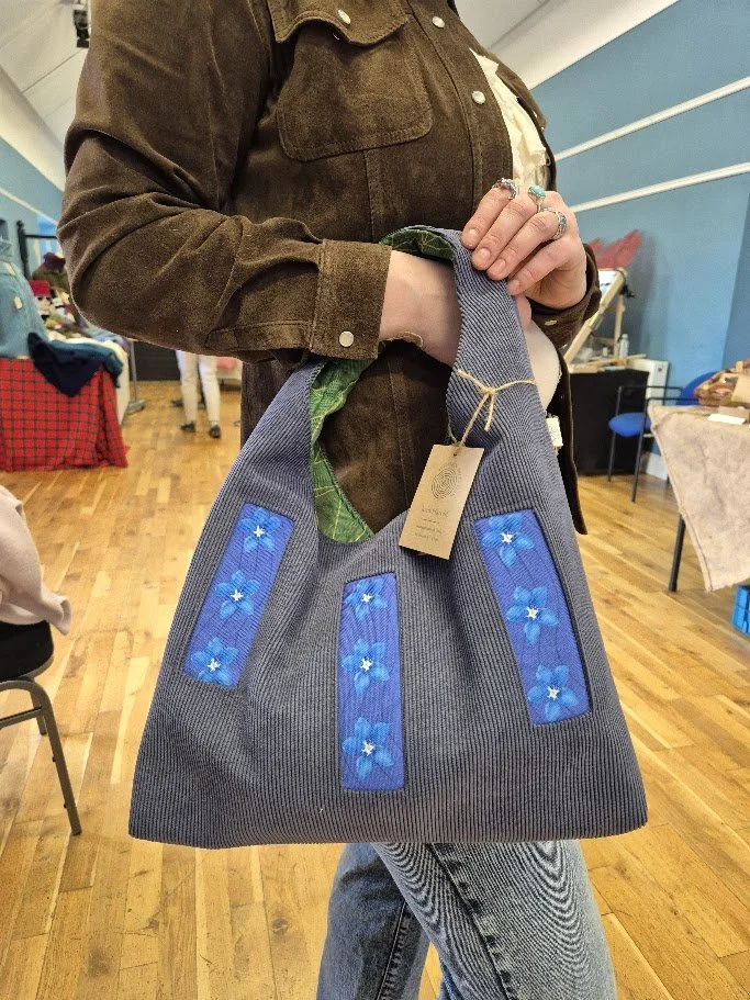 Person holding a large gray bag with blue flower designs on it, inside a room with wooden floors and tables.