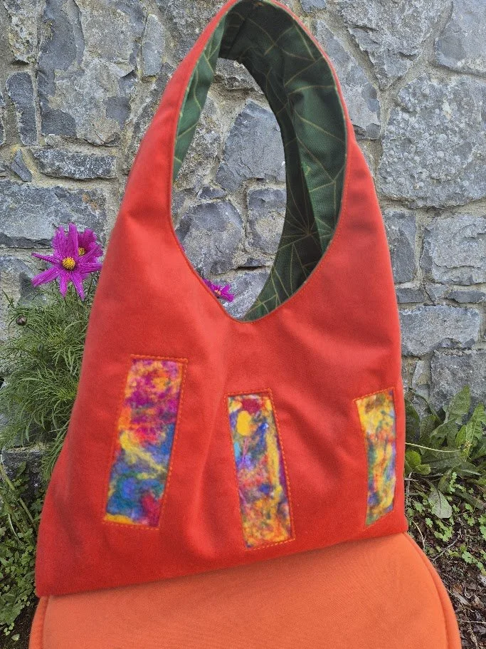 Red fabric tote bag with colorful abstract patches hanging against a stone wall with purple flowers nearby.