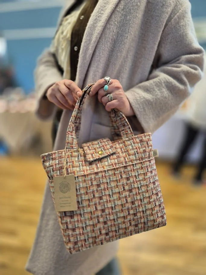 A person in a beige coat holding a woven fabric purse with a handle and a snap button, standing indoors with a blurred background.