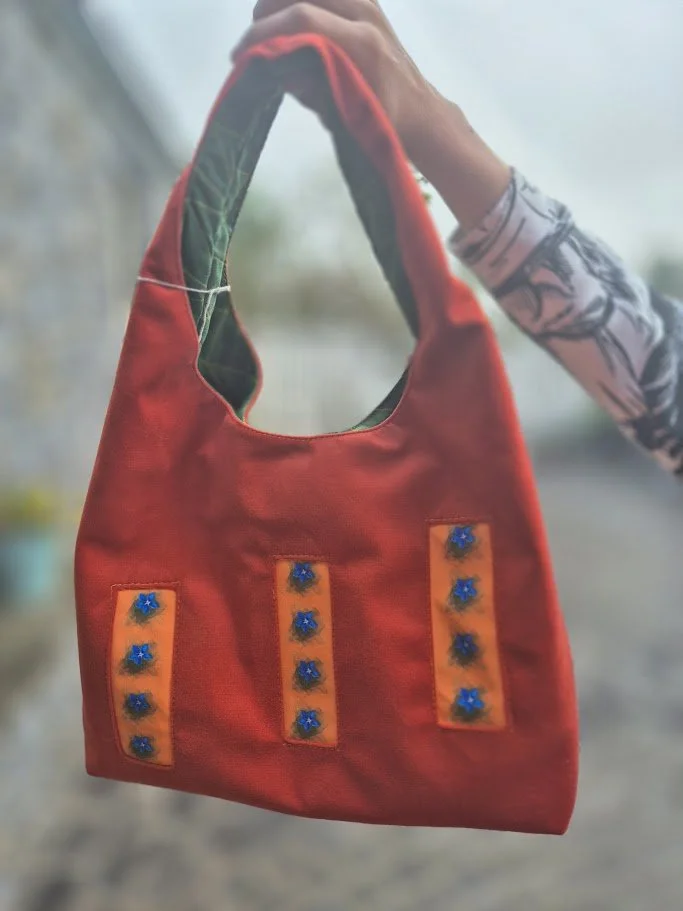 A handmade fabric bag with vertical orange patches decorated with blue beads, being held by a person wearing a patterned long sleeve shirt.