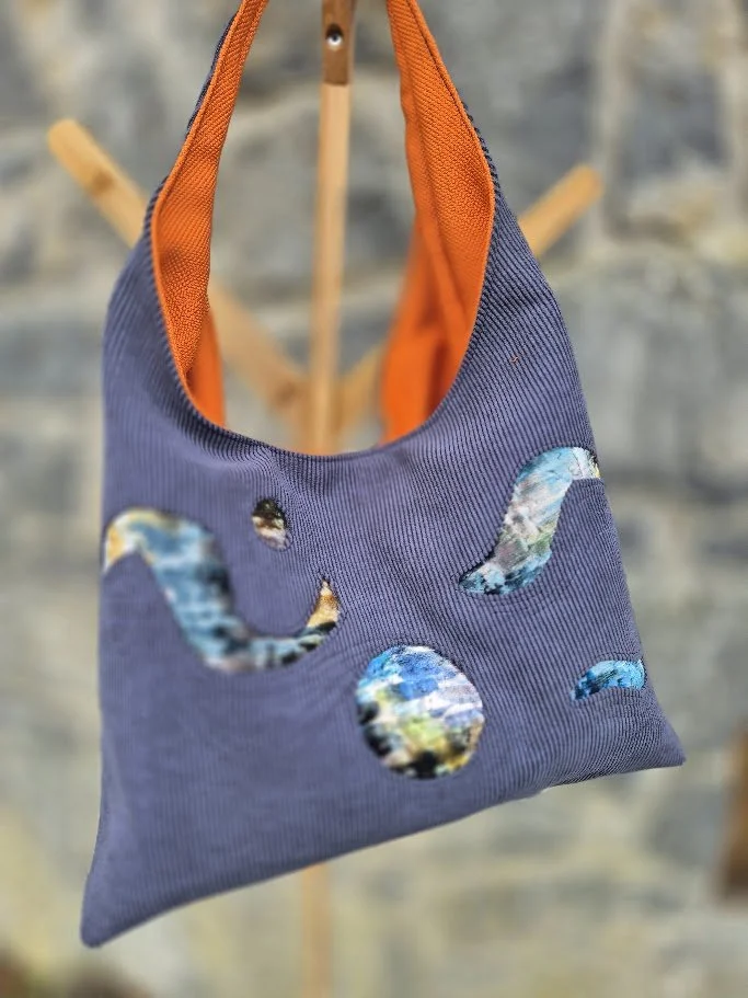 A fabric tote bag with blue and orange colors, decorated with cut-out shapes resembling moons, clouds, and raindrops, hanging on a wooden stand against a gray brick wall.